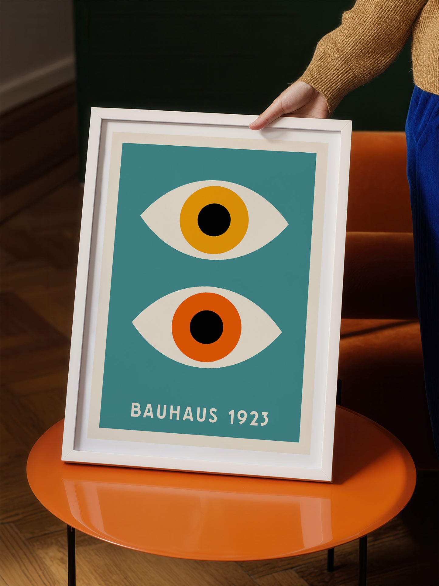 A person is holding up a framed poster with a minimalist design featuring two stylized eyes on a teal background.