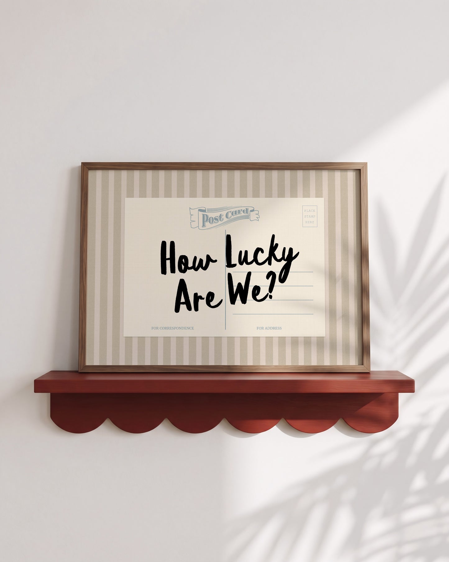A framed postcard with the text "How Lucky Are We?" displayed on it, placed on a wooden shelf against a white wall.