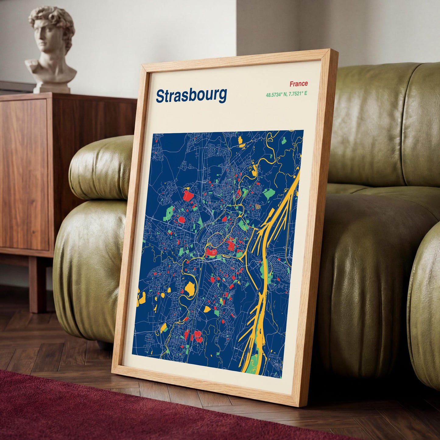 A framed map of Strasbourg, France, is displayed on a wooden floor in front of a green leather couch.