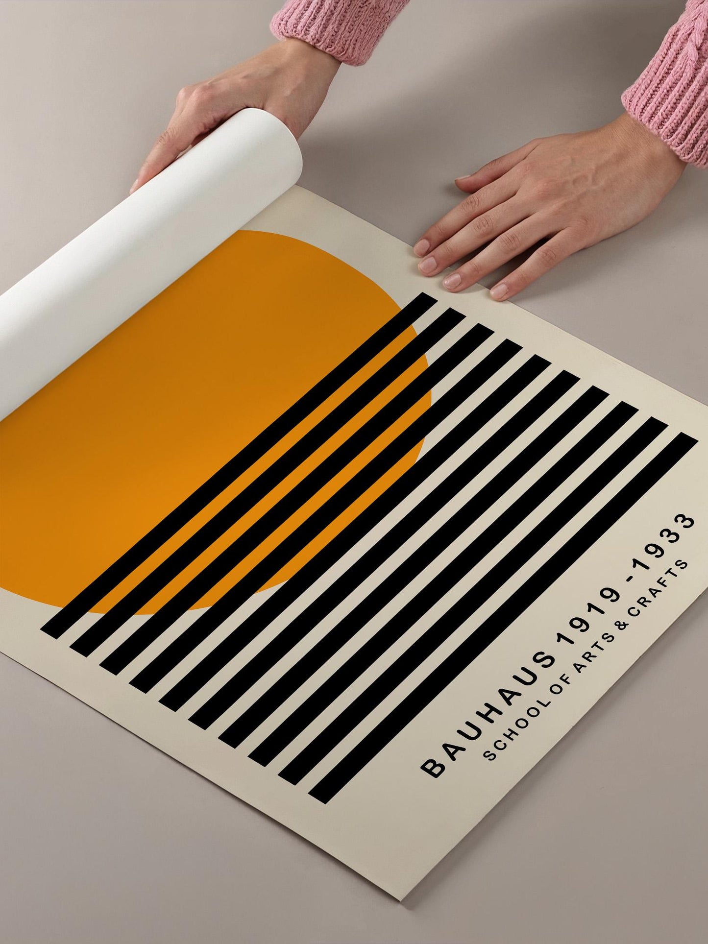 A person's hands are holding a roll of paper, which appears to be a poster or artwork featuring a bold, geometric design with black and orange stripes and a large orange circle.