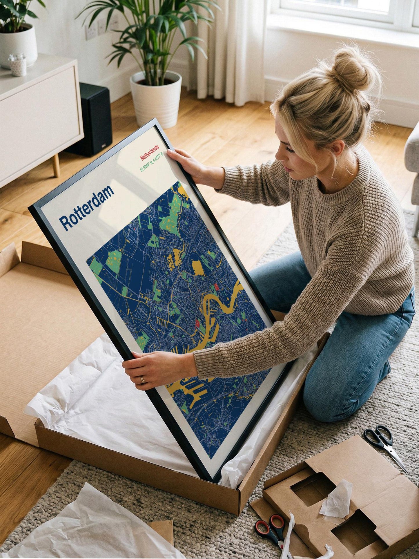 A woman is kneeling on the floor and holding a framed map of Rotterdam, a city in the Netherlands.