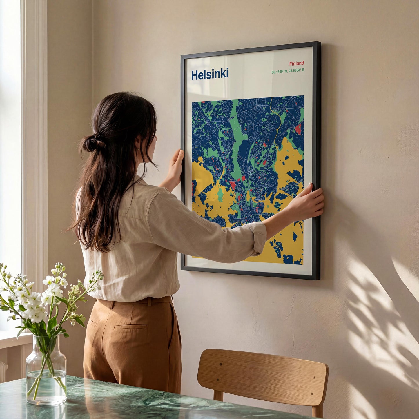 A woman is hanging a framed map of Helsinki, Finland, on a wall in a room with a dining table and a vase of flowers.