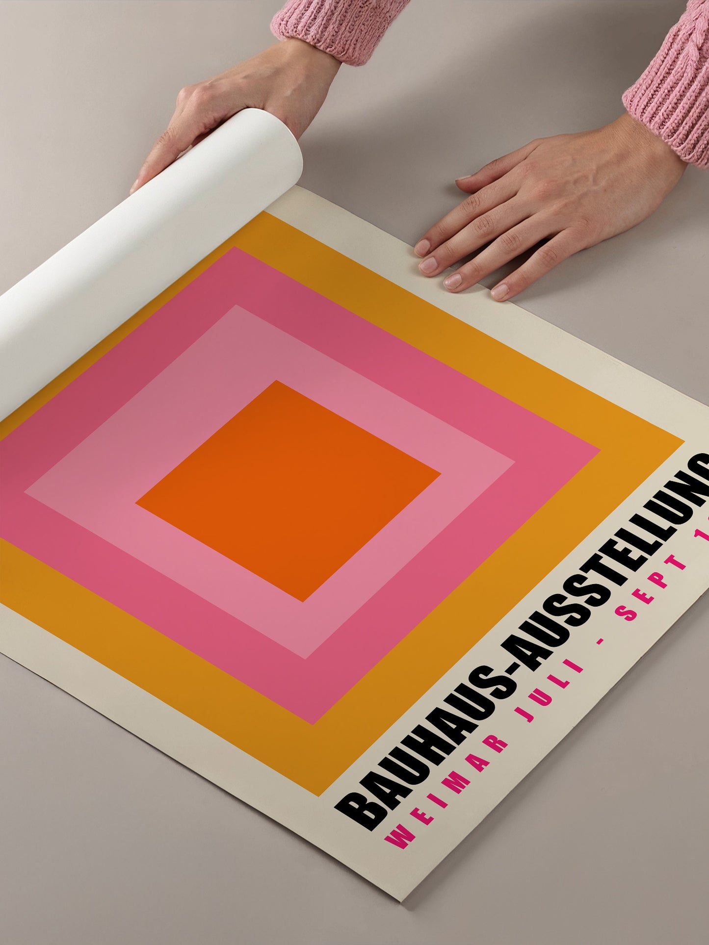 A person's hands are holding a roll of paper, which appears to be a poster or flyer for an event. The poster has a bold, geometric design with a large orange square in the center, surrounded by a gradient of pink and yellow squares. The text on the poster indicates that this is an exhibition of Bauhaus art, featuring works by Jümar Juli.