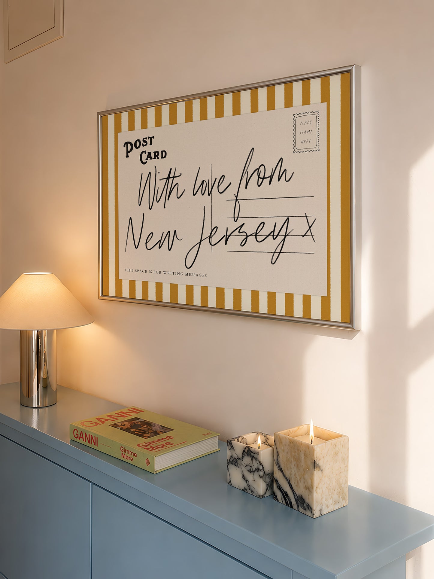 a framed postcard with a handwritten message, a lamp, a book, and a marble candle holder on a blue dresser.