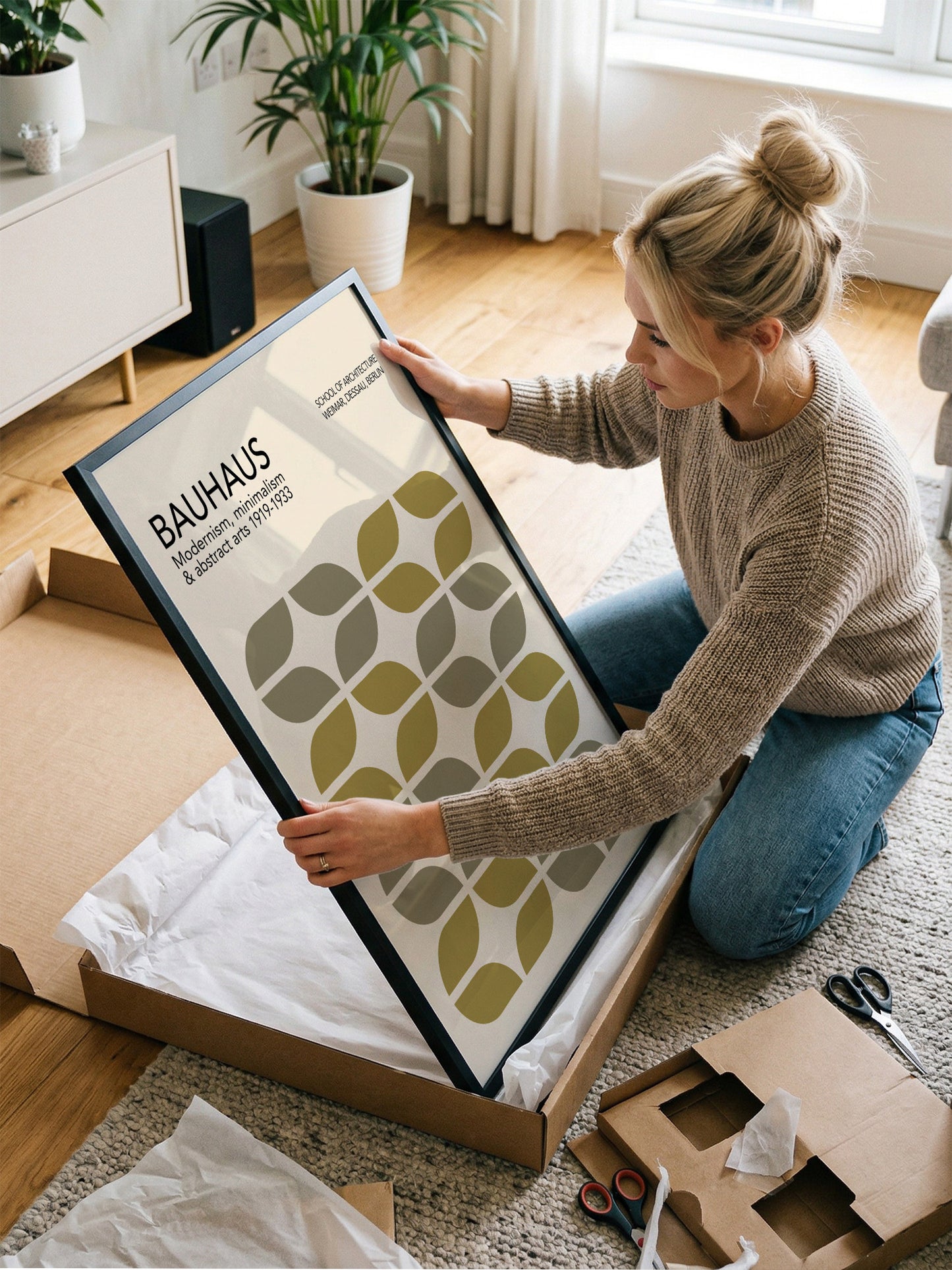 A woman is kneeling on the floor and holding a framed poster with a geometric pattern.