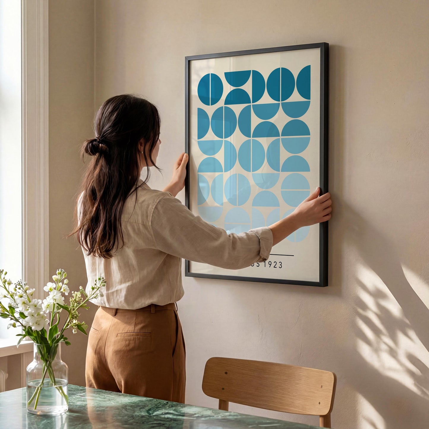 A woman is hanging a framed artwork on a wall in a room with a dining table and chair visible.