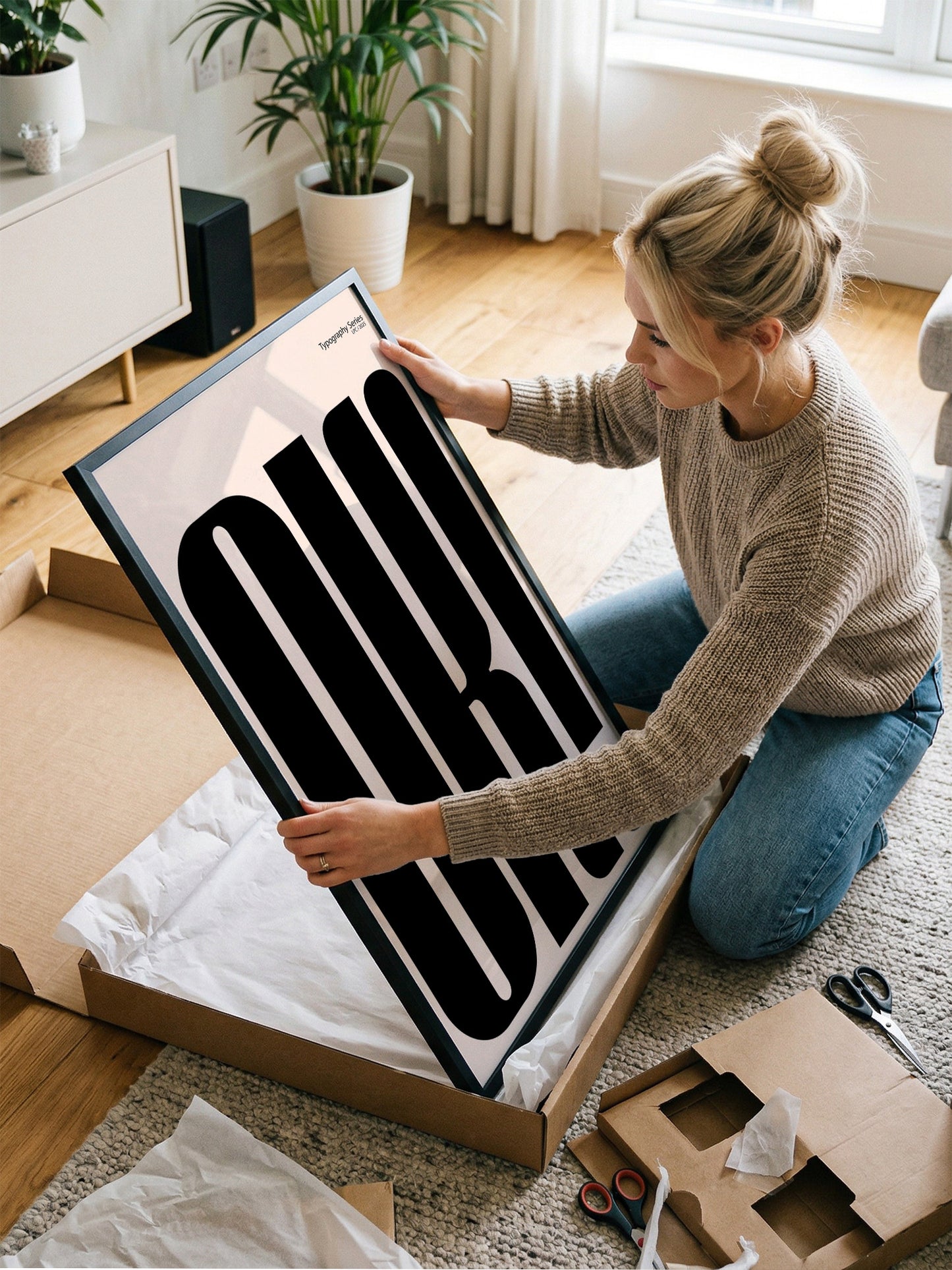 A woman is kneeling on the floor and carefully handling a large framed print, which appears to be a piece of artwork or a poster.