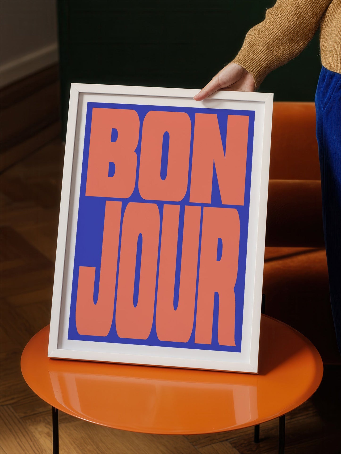 A person is holding up a framed poster with the text "BON JOUR" in large, bold letters.
