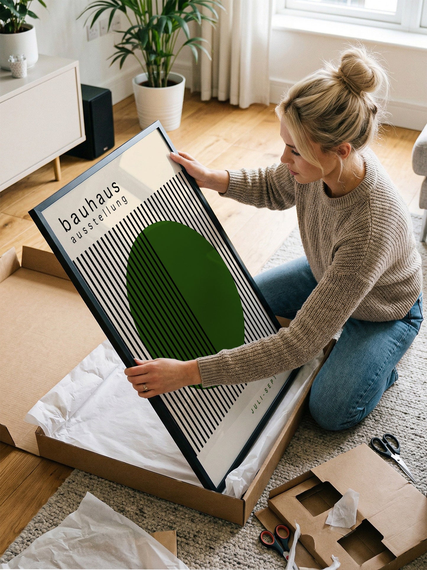 A woman is kneeling on the floor and carefully opening a large cardboard box, which contains a framed poster with a green circle and black stripes.