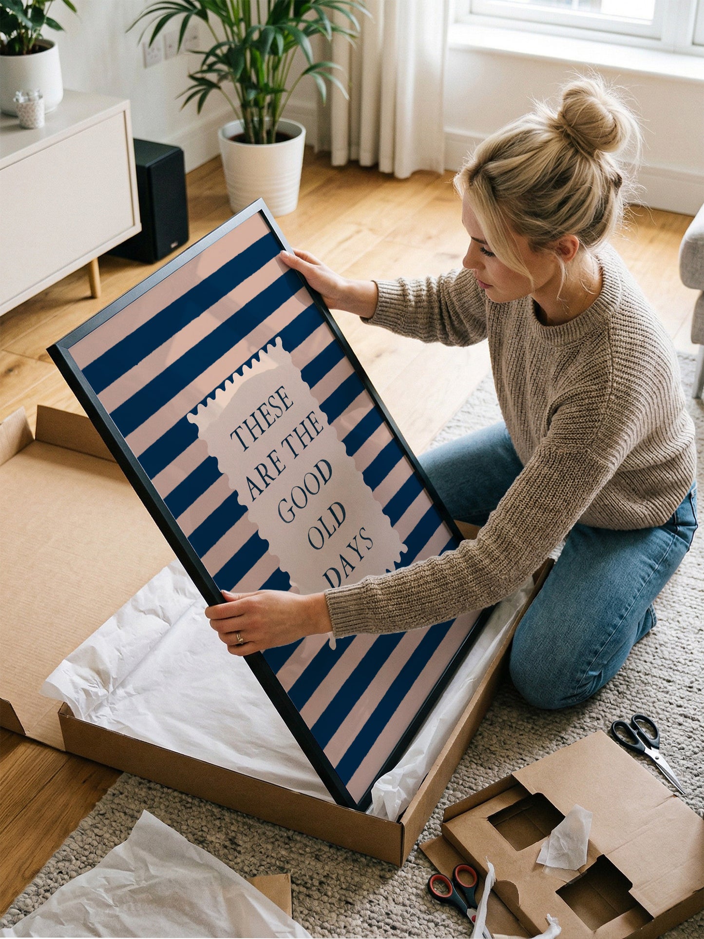 A woman is kneeling on the floor and holding a framed poster with the text "These are the good old days" on it.