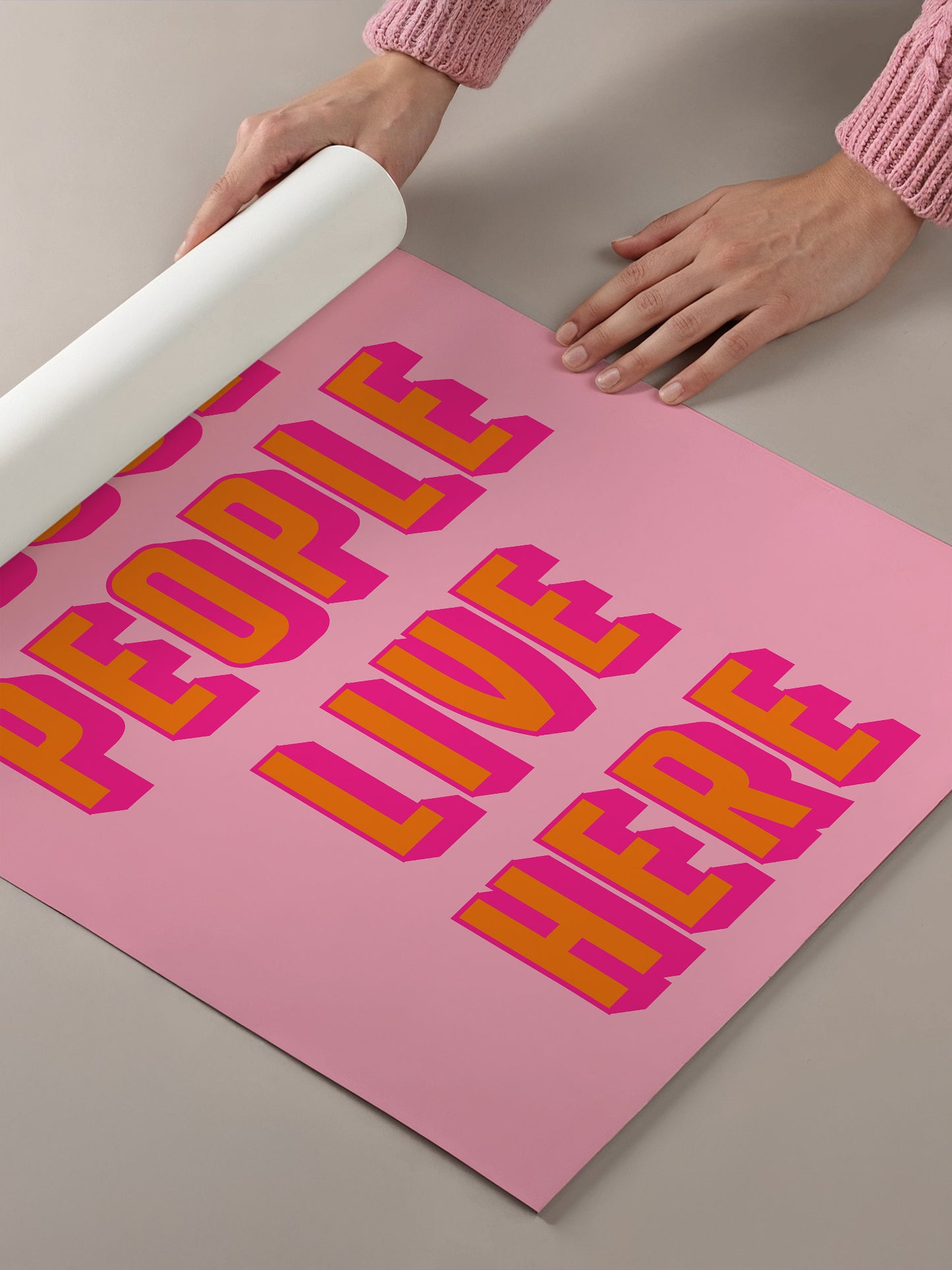 A person's hands are holding a roll of paper, which is placed on top of a pink poster with large, bold text that reads "PEOPLE LIVE HERE".