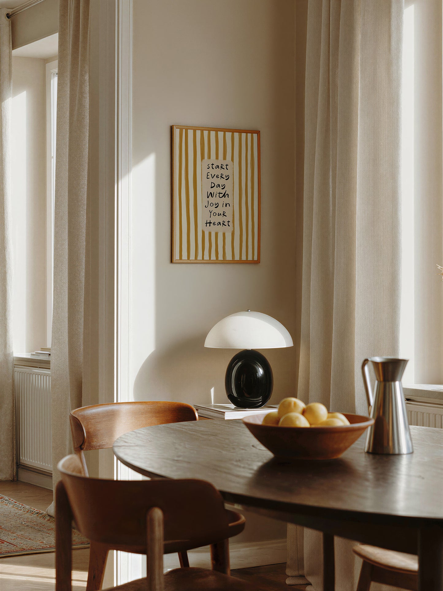 a cozy dining area with a wooden table, two chairs, and a lamp. A framed quote is hanging on the wall above the table, and a bowl of fruit is placed on the table.