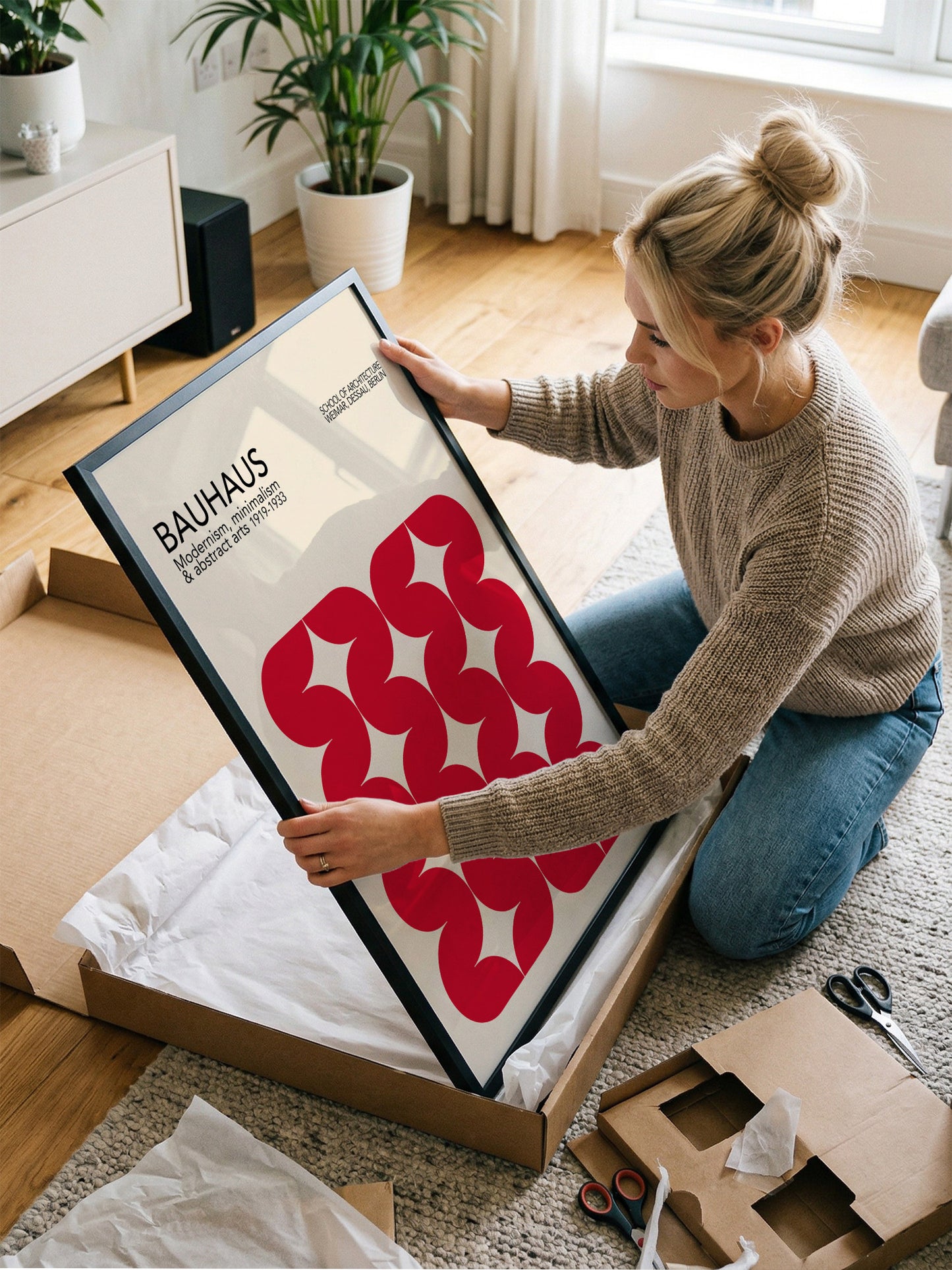 A woman is kneeling on the floor and holding a framed poster with a red and white design.