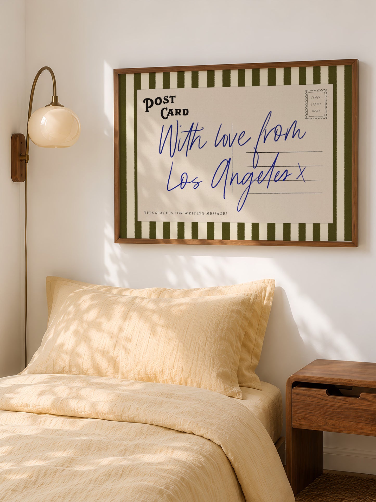 a bedroom with a bed, a nightstand, and a framed postcard hanging on the wall. The postcard has a handwritten message that reads "With love from Los Angeles x" and is framed with a green and white striped border.
