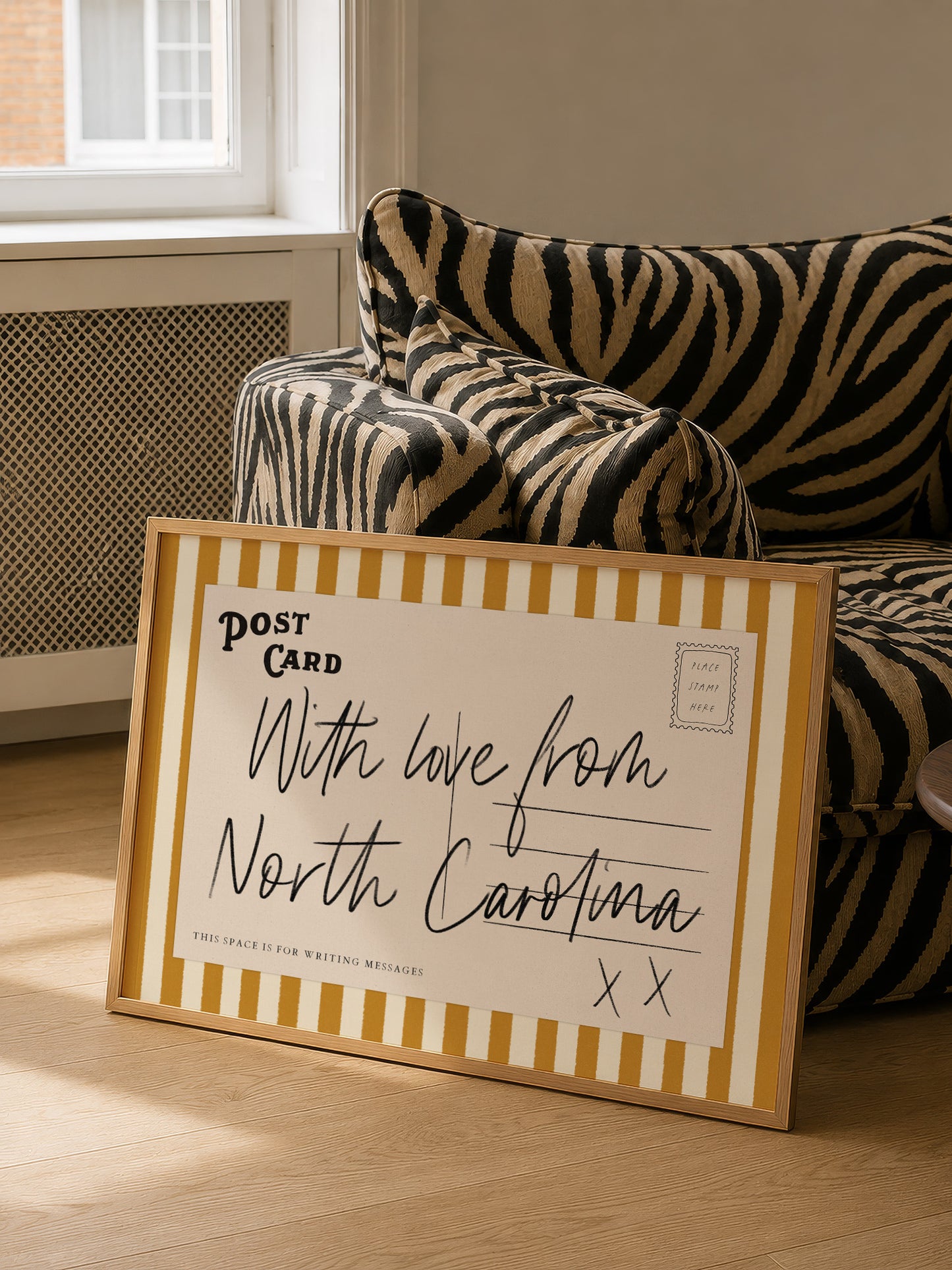 A framed postcard with a handwritten message is placed on a couch in a room with a window.