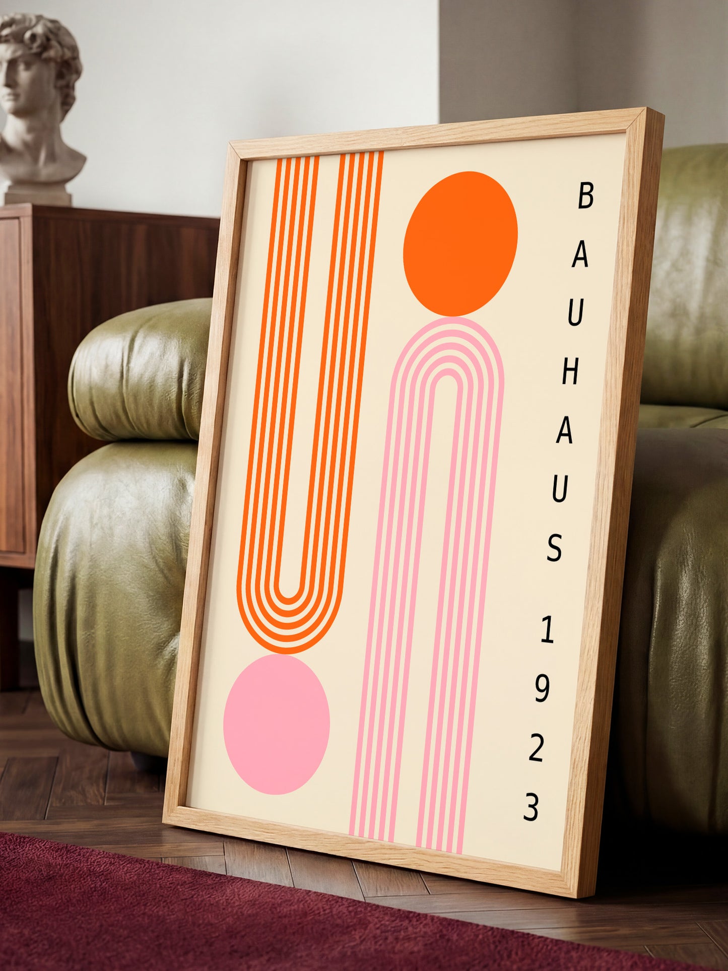 a framed poster with abstract geometric designs in orange and pink colors, placed on a wooden floor in front of a green leather couch.