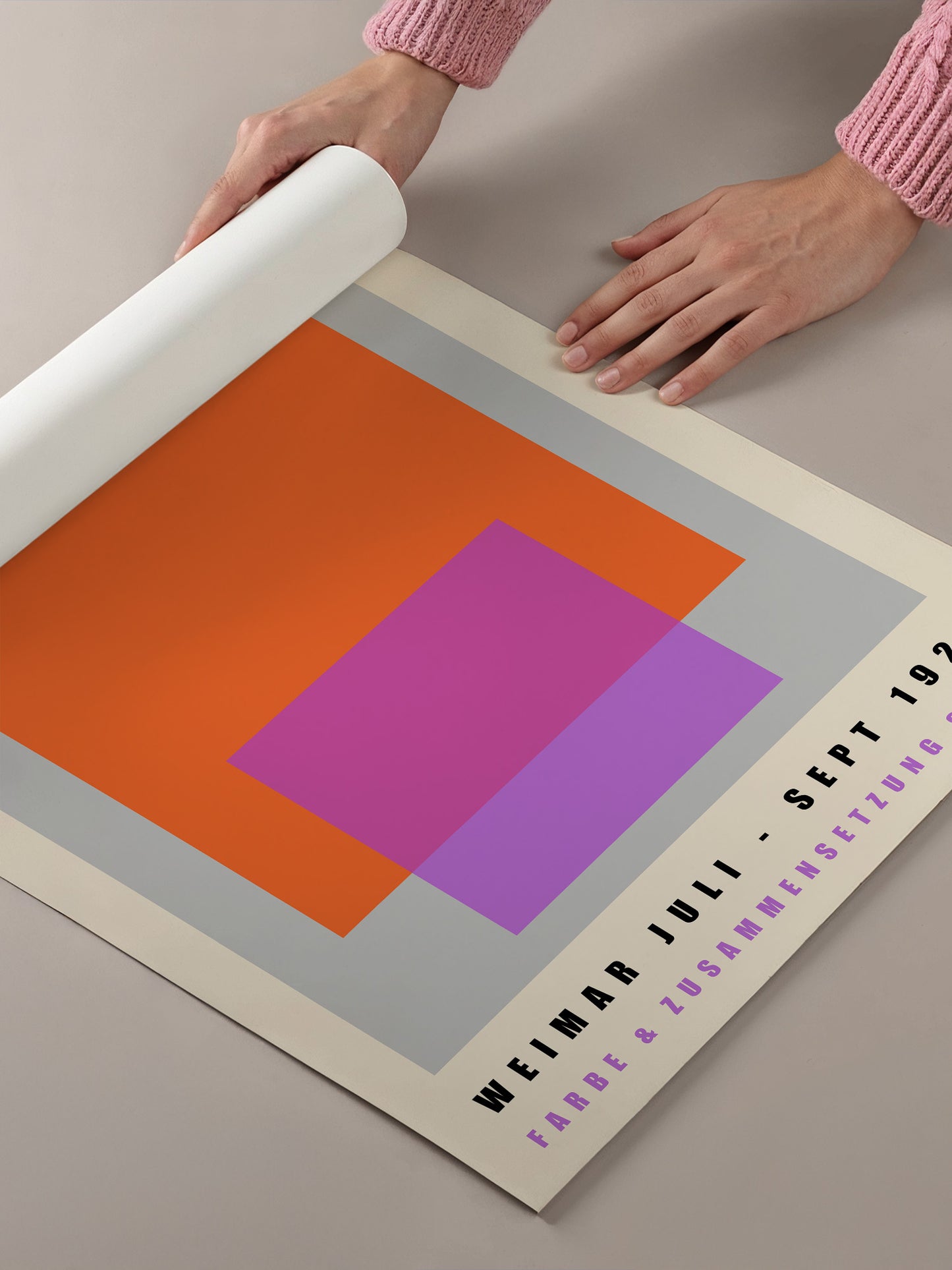 A person's hands are holding a roll of paper, which appears to be a poster or flyer with a geometric design featuring orange, pink, and purple shapes.