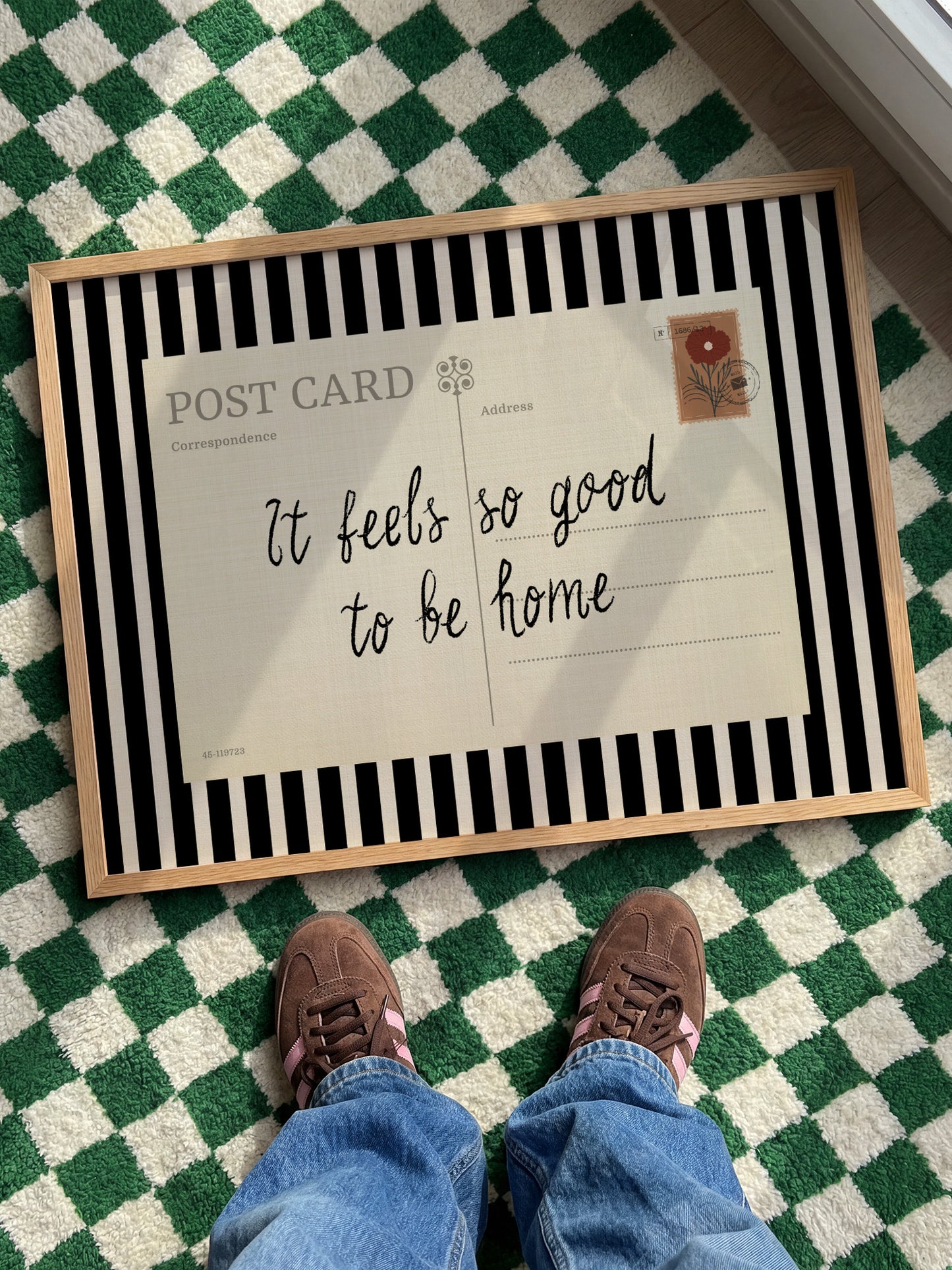 A person's feet standing on a checkered green and white rug, with a framed postcard underneath that reads "It feels so good to be home".
