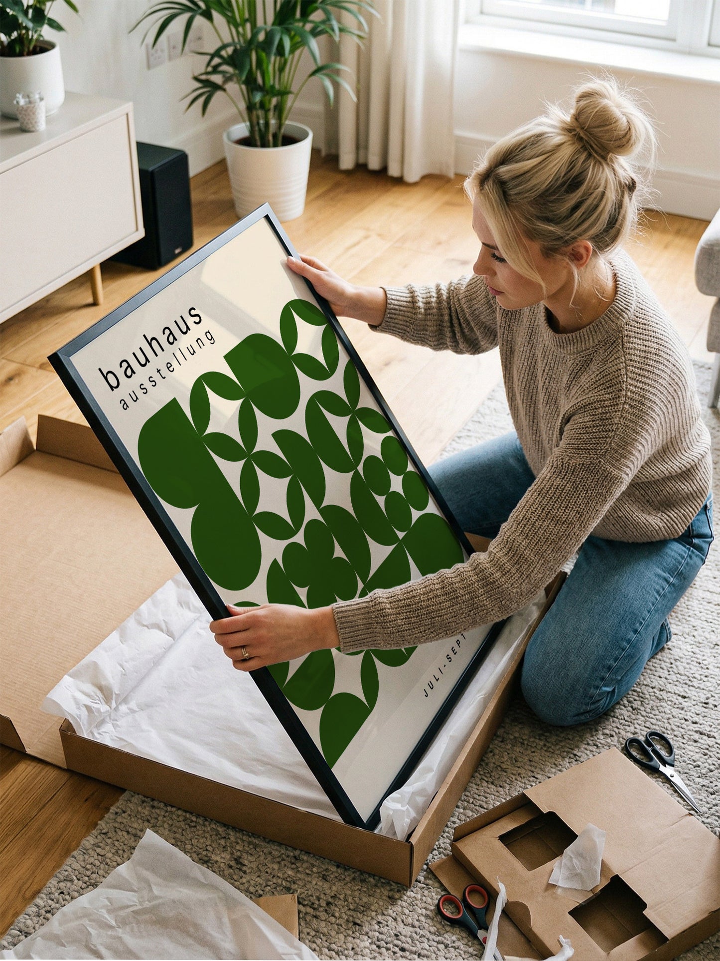 A woman is kneeling on the floor and holding a framed poster with a green and white design.