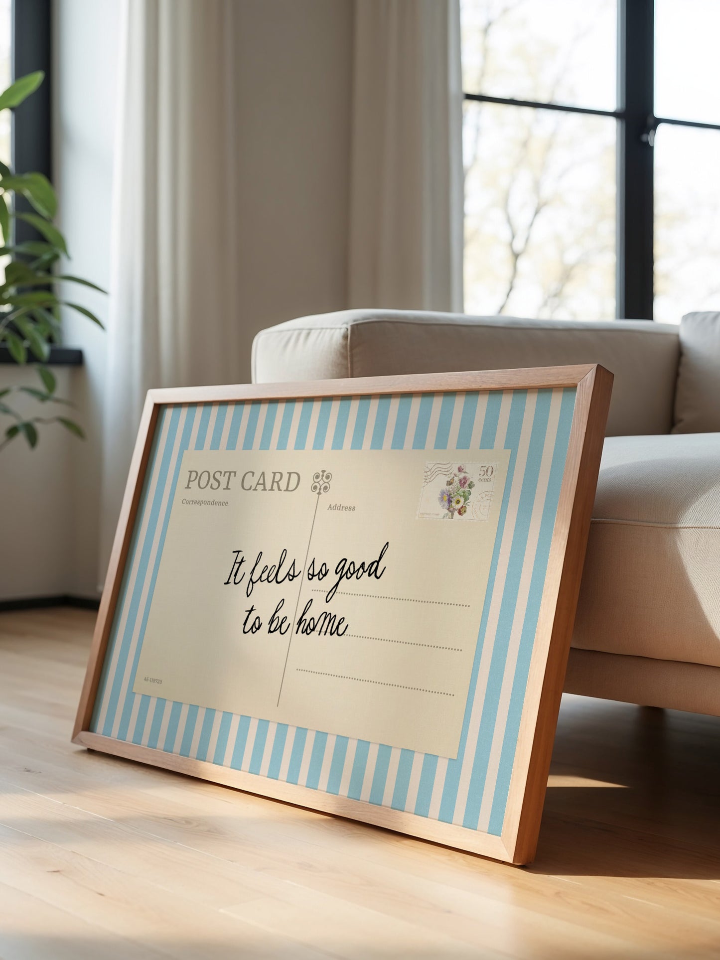 A framed postcard with a handwritten message sits on a wooden floor in a room with a window and a couch.
