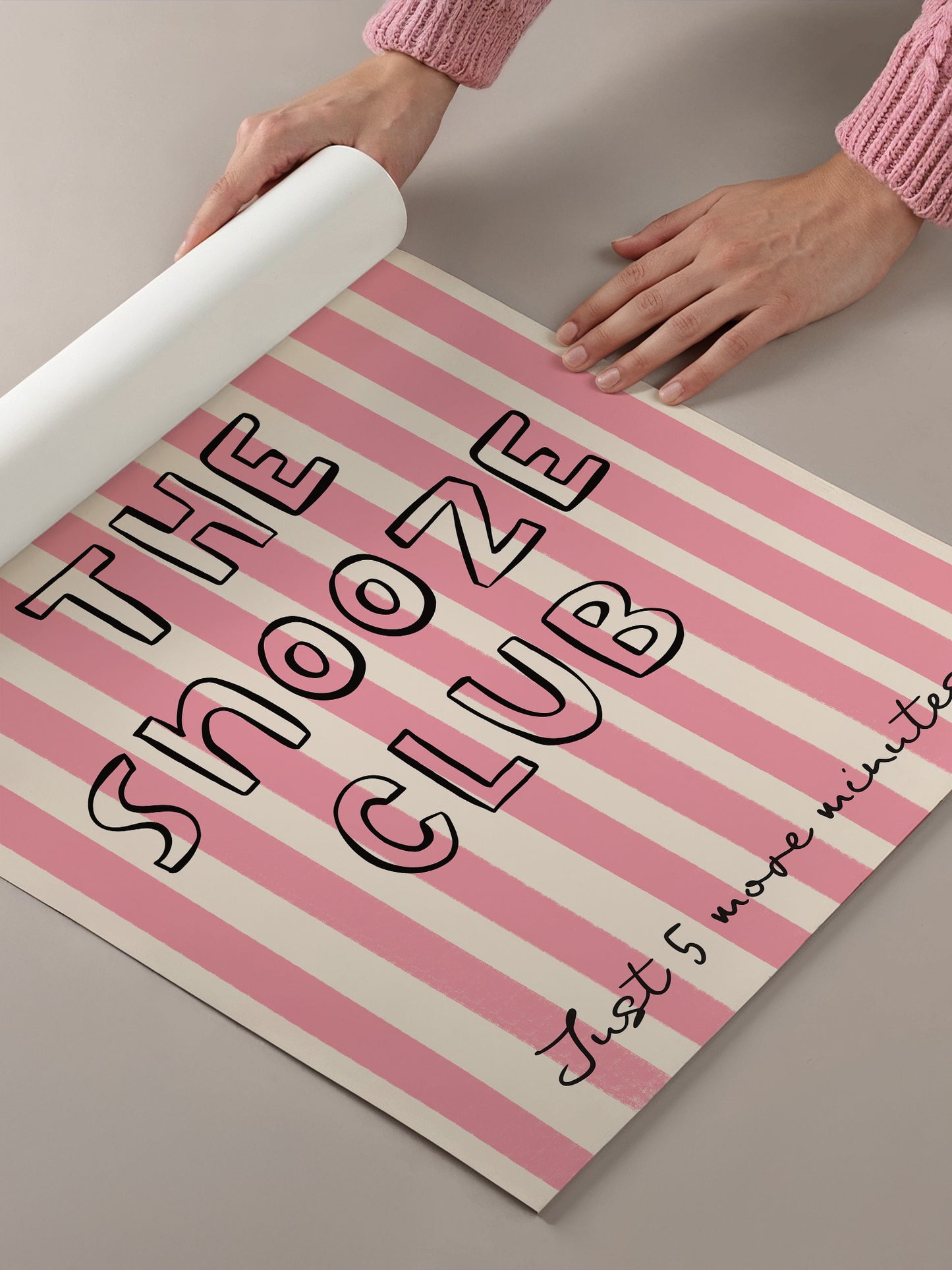 A person's hands are holding a roll of paper with the text "THE SNOOZE CLUB" printed on it, which is a pink and white striped background.