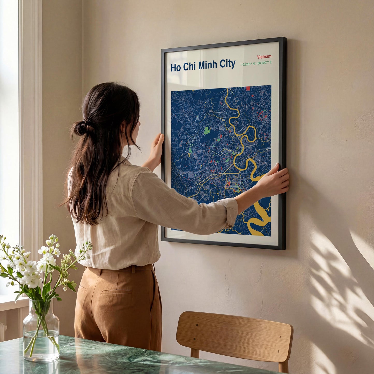A woman is hanging a framed map of Ho Chi Minh City on a wall in a room with a dining table and a vase of flowers.