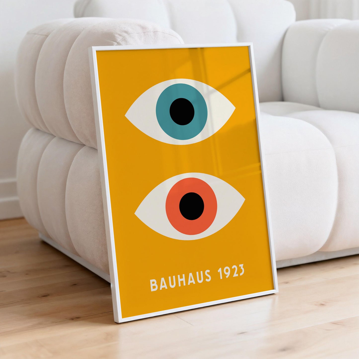 A framed poster with a yellow background and two stylized eyes, one blue and one orange, sitting on a white couch.
