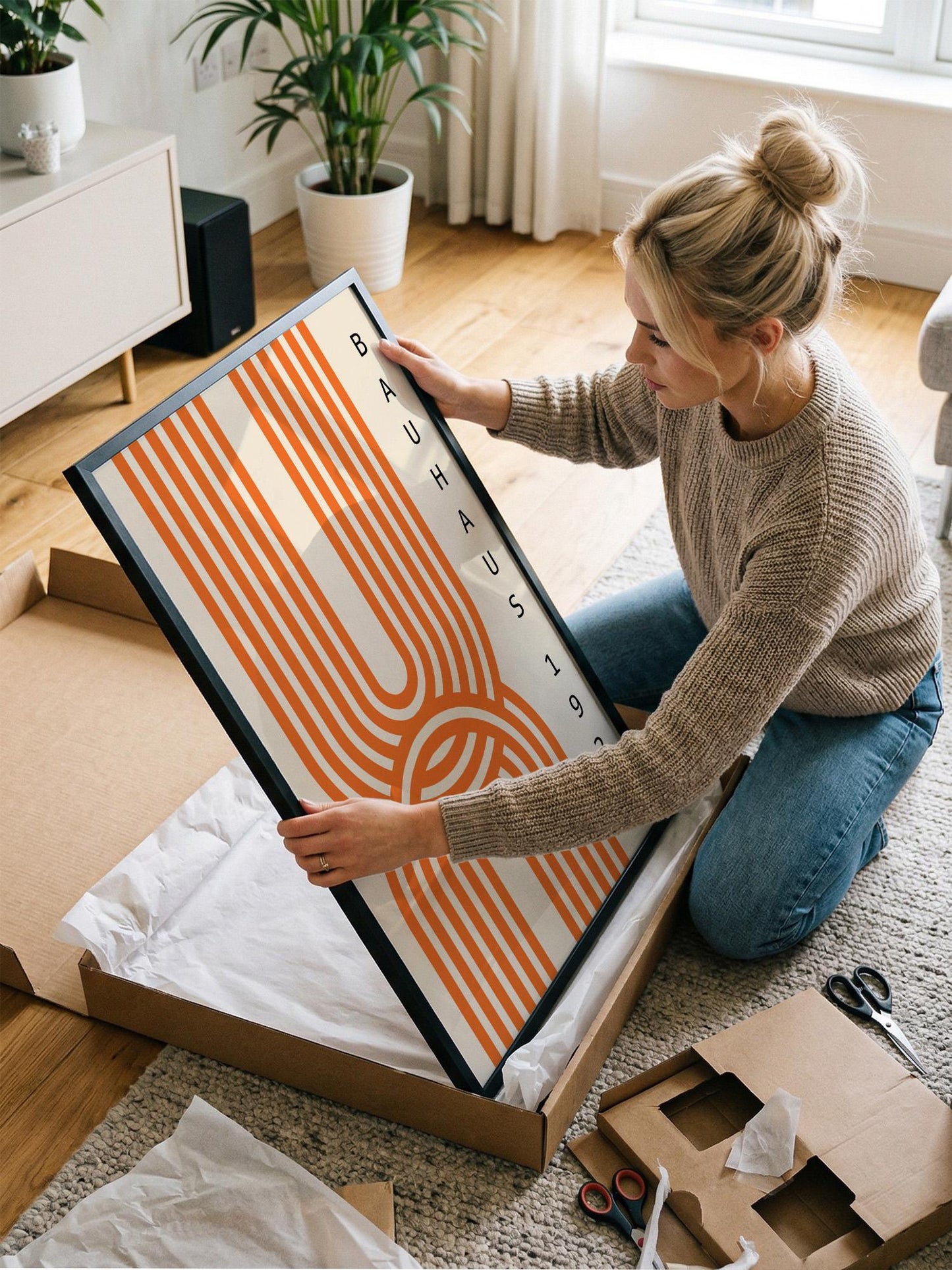 A woman is kneeling on the floor and carefully opening a large cardboard box, which contains a framed piece of art with a striking orange and white striped design.