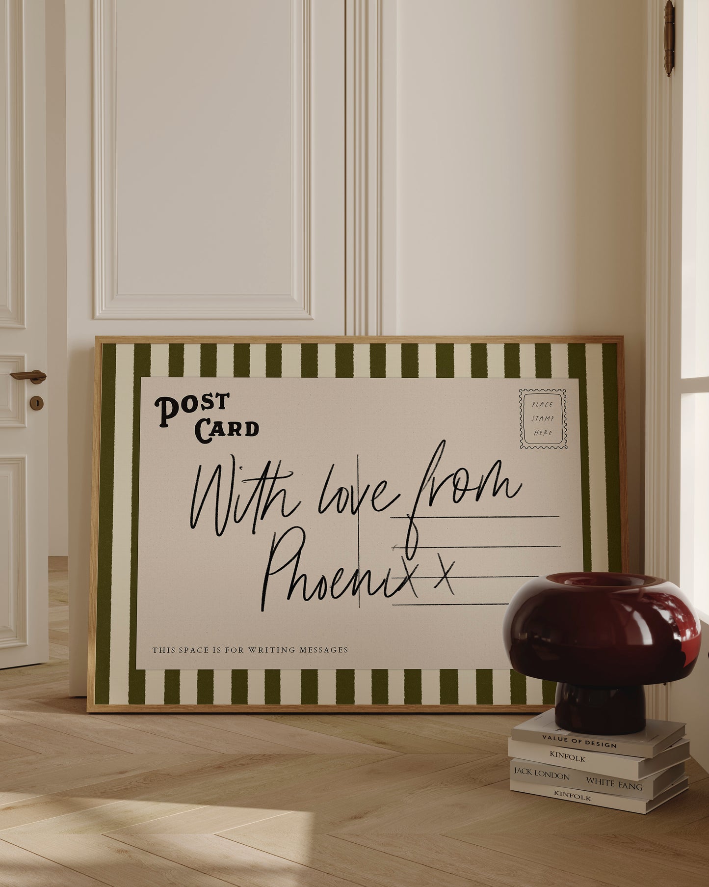 a large framed postcard with a handwritten message, placed on a wooden floor next to a stack of books and a lamp.