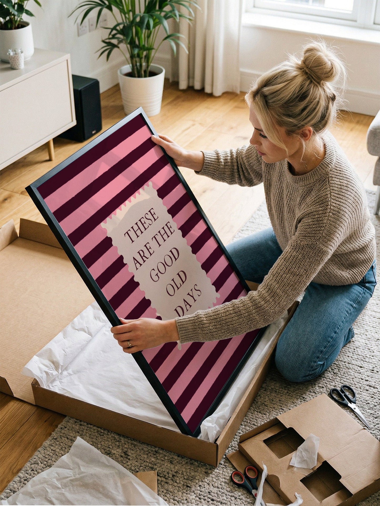A woman is kneeling on the floor and holding a large framed poster with the text "These are the good old days" on it.