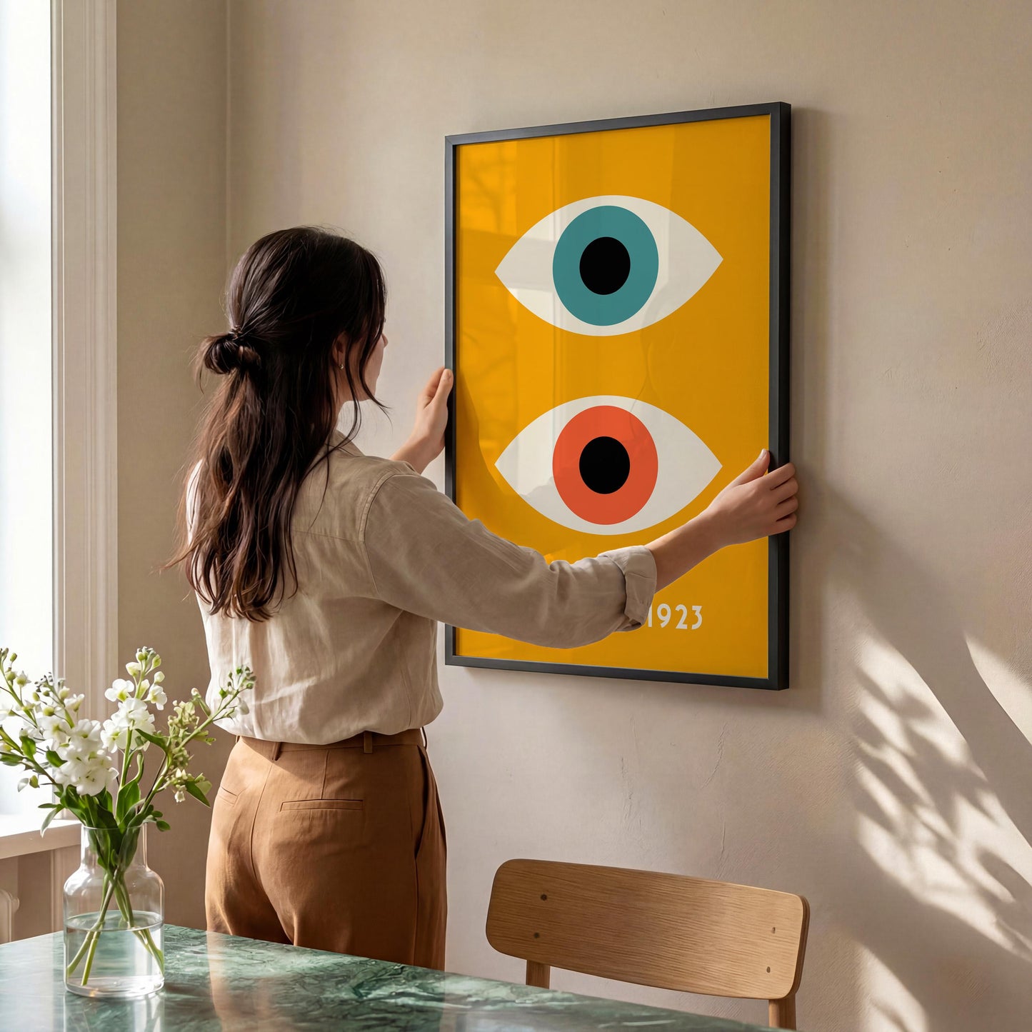A woman is hanging a framed poster on a wall, which features a stylized design of two eyes.
