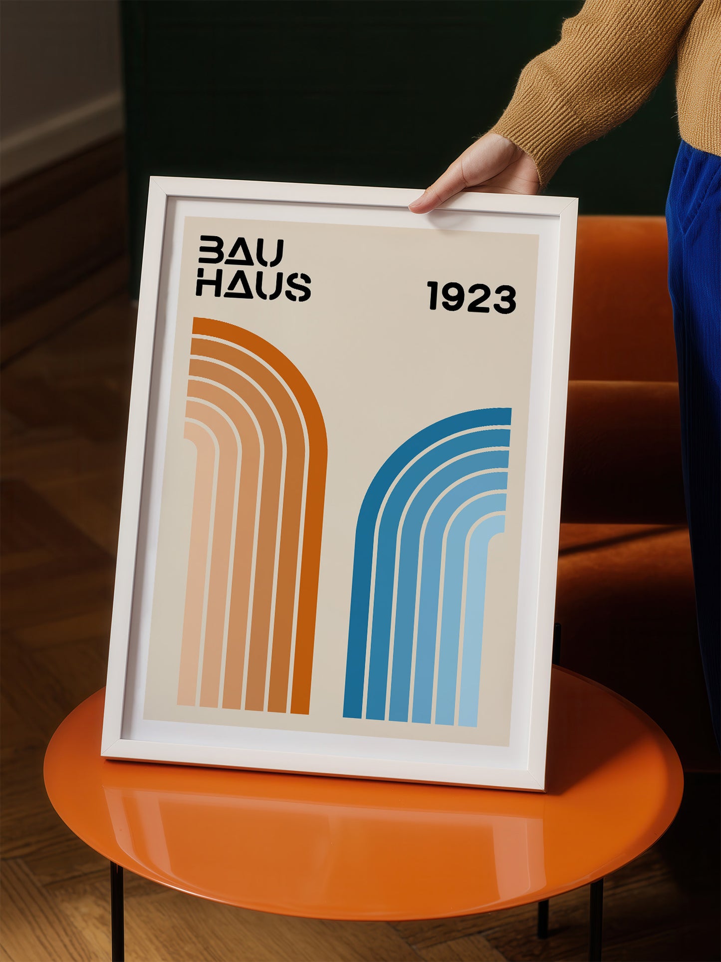A person is holding a framed poster with the text "BAU HAUS 1923" and two abstract geometric designs in orange and blue.