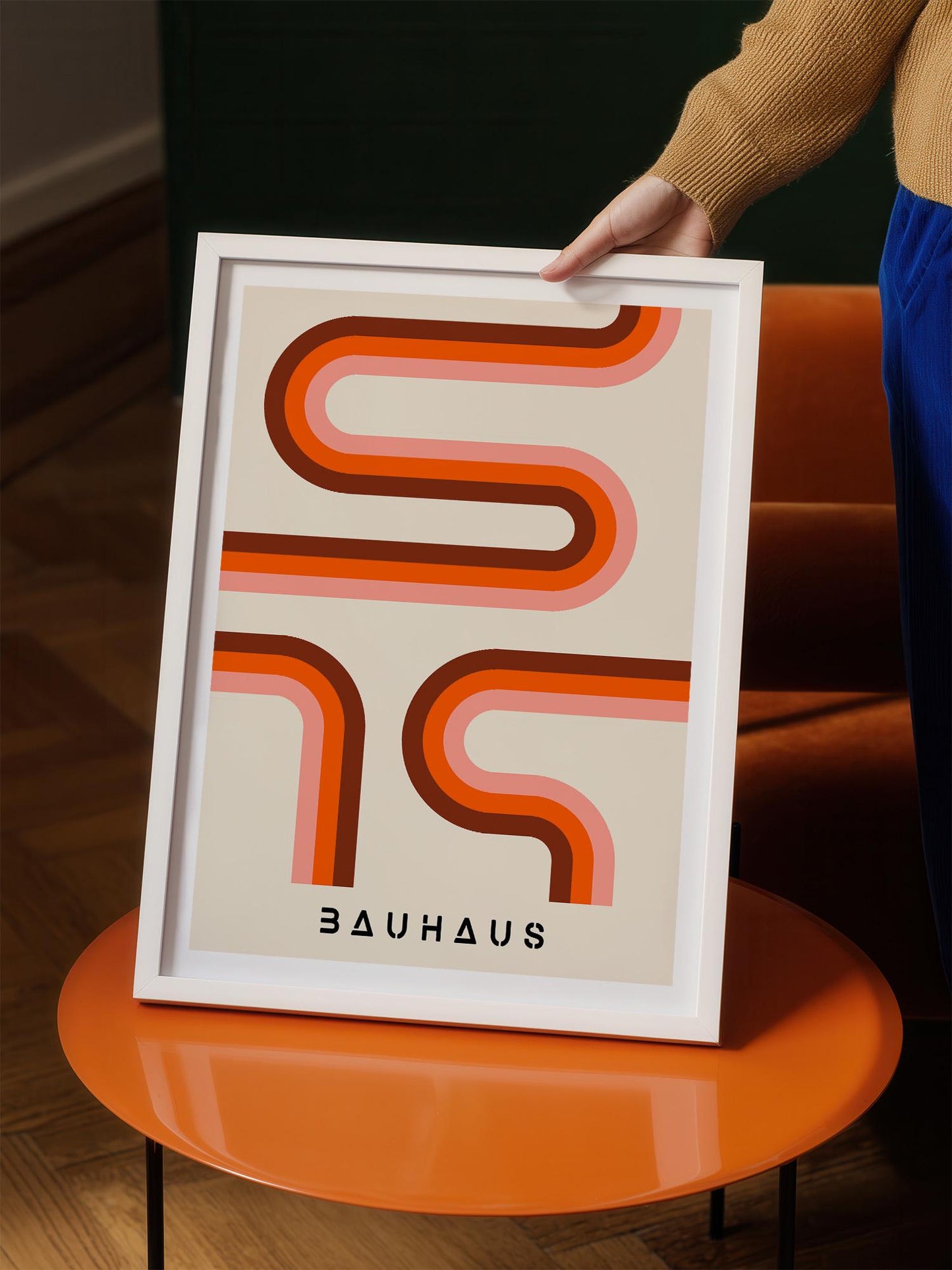 A person is holding up a framed poster with a minimalist design featuring a repeating pattern of curved lines in shades of orange and pink.