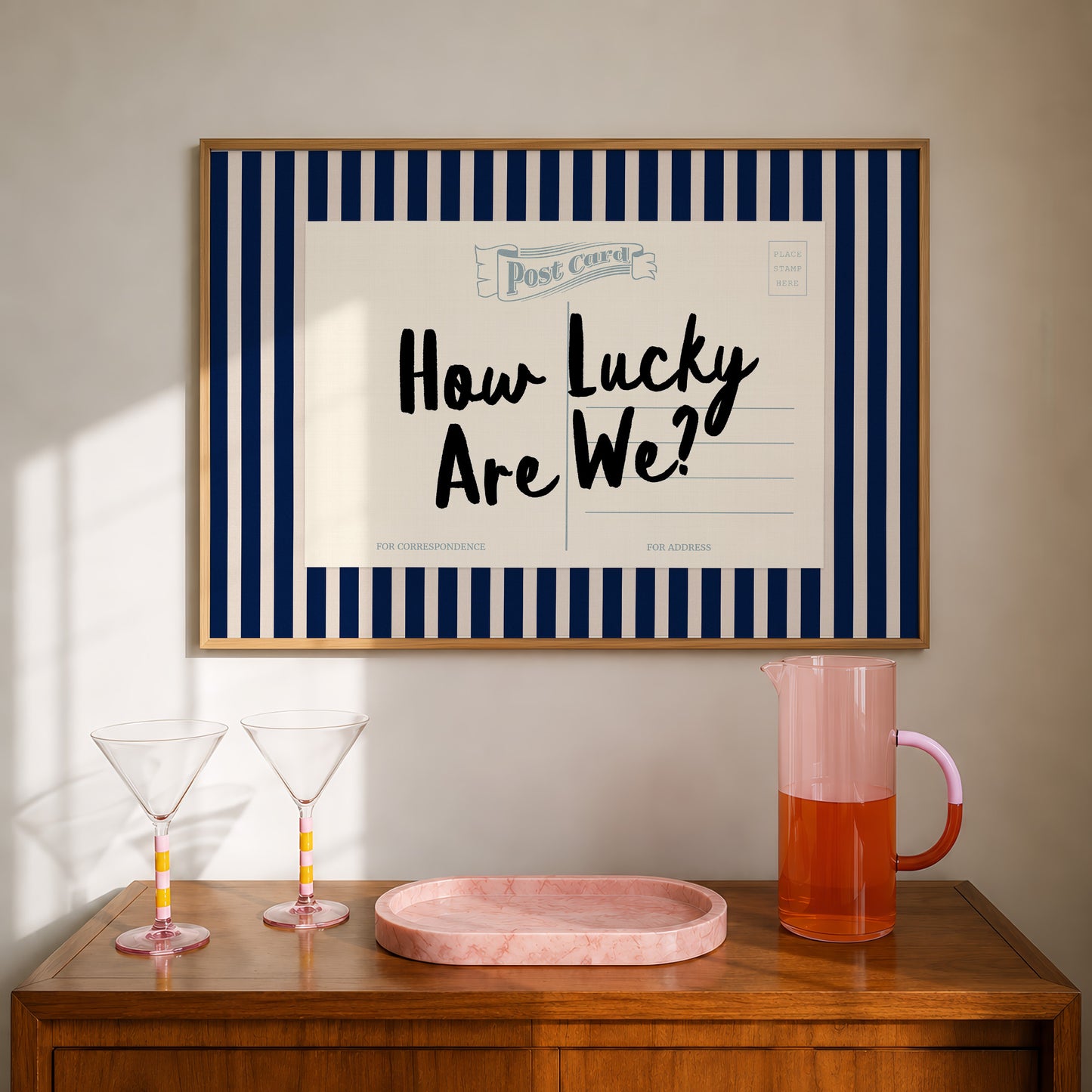 a framed poster with the text "How Lucky Are We?" hanging on a wall above a wooden table with two martini glasses and a pink pitcher.