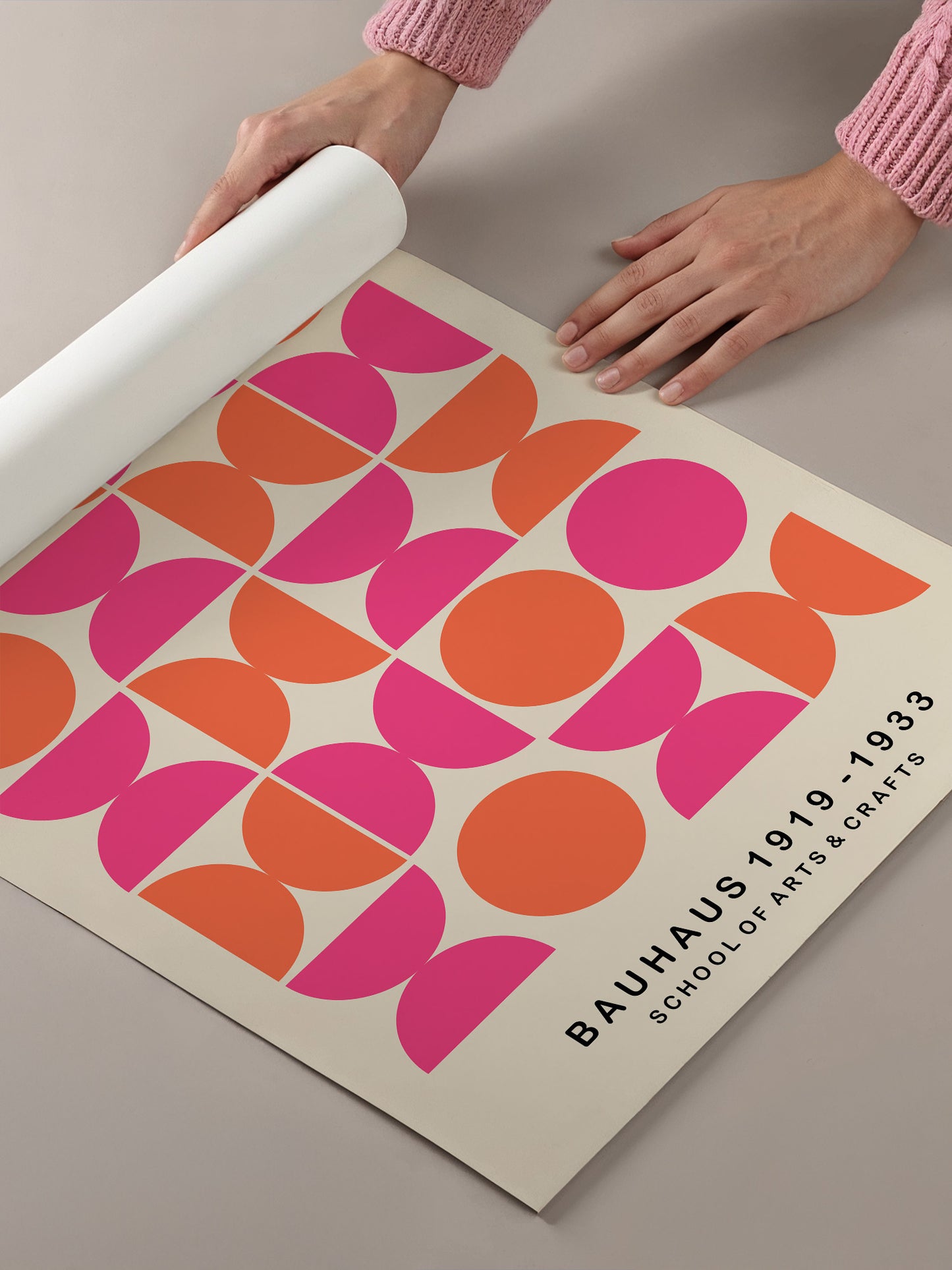 A person's hands are holding a roll of paper, which appears to be a poster or artwork featuring a geometric design with circles and lines in various shades of pink and orange.
