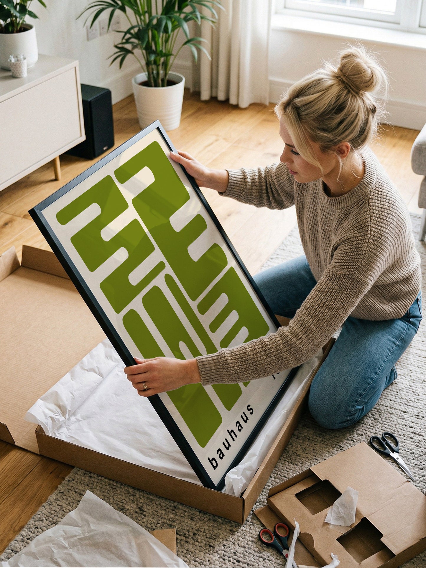 A woman is kneeling on the floor and holding a large framed poster with a green and white design.