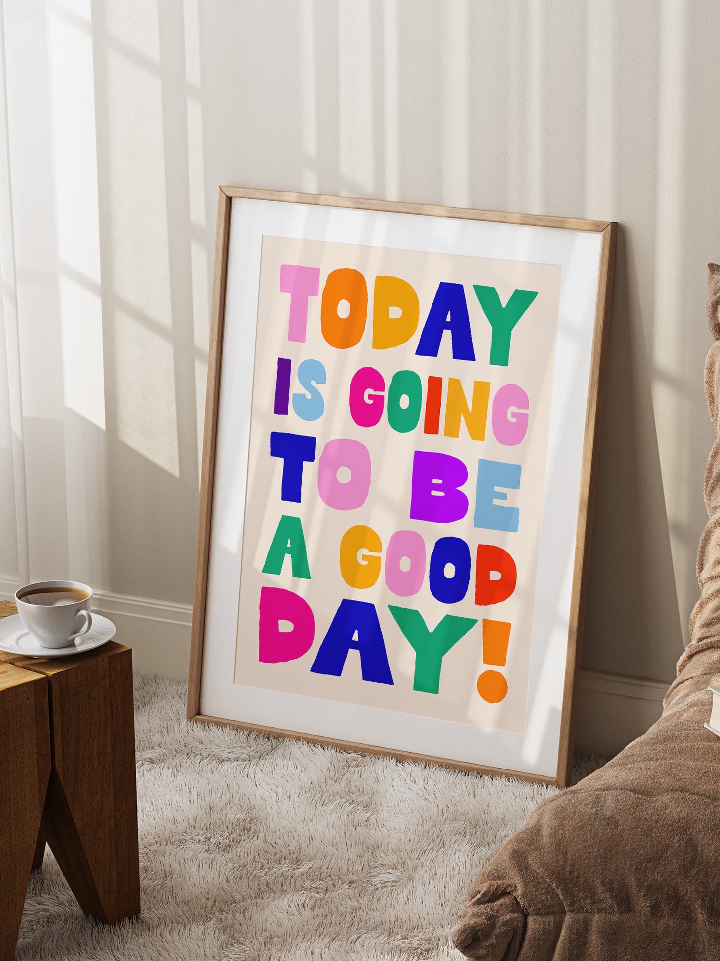 A framed poster with the text "TODAY IS GOING TO BE A GOOD DAY!" is displayed on a wall in a room with a wooden table and a coffee cup nearby.