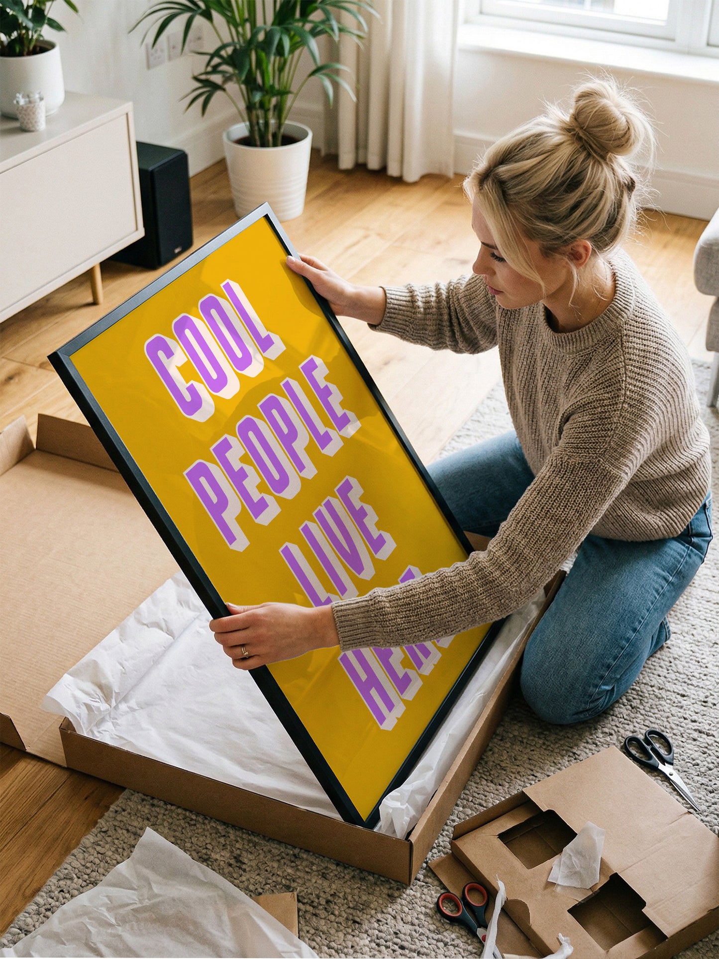 A woman is kneeling on the floor and holding a large framed poster with the text "Cool people live here" on it.