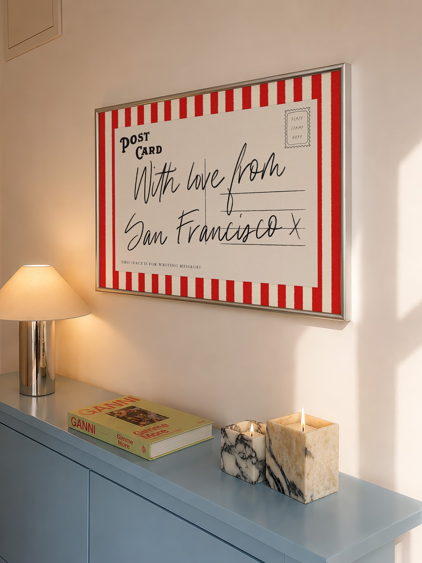 a framed postcard with a handwritten message, a lamp, a book, and a marble candle holder on a blue dresser.