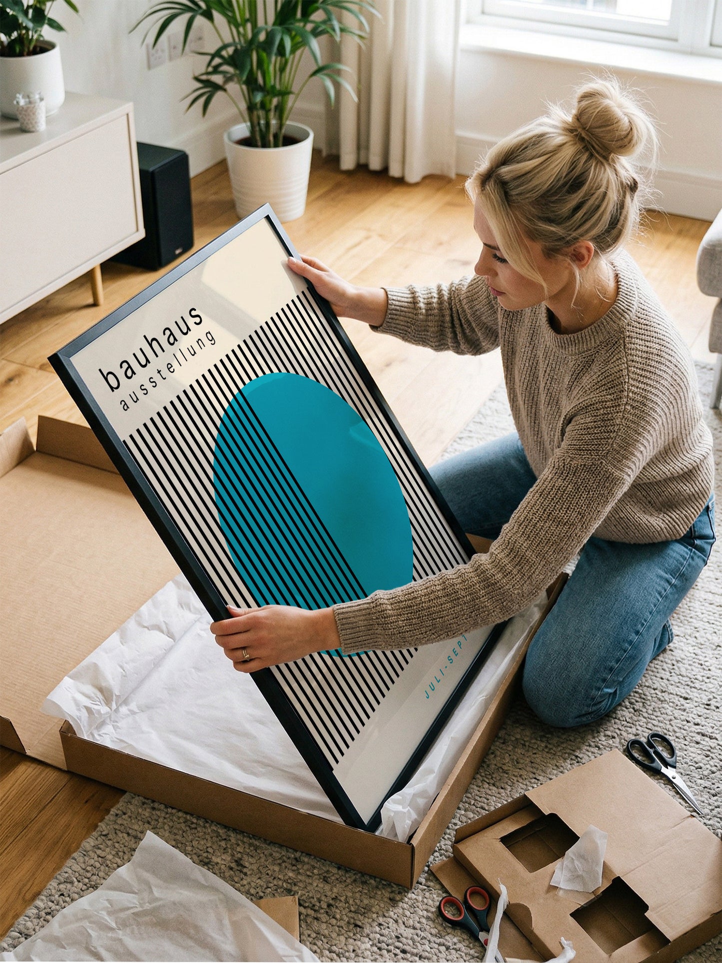 A woman is kneeling on the floor and carefully opening a large cardboard box, which contains a framed poster with a striking blue and black design.