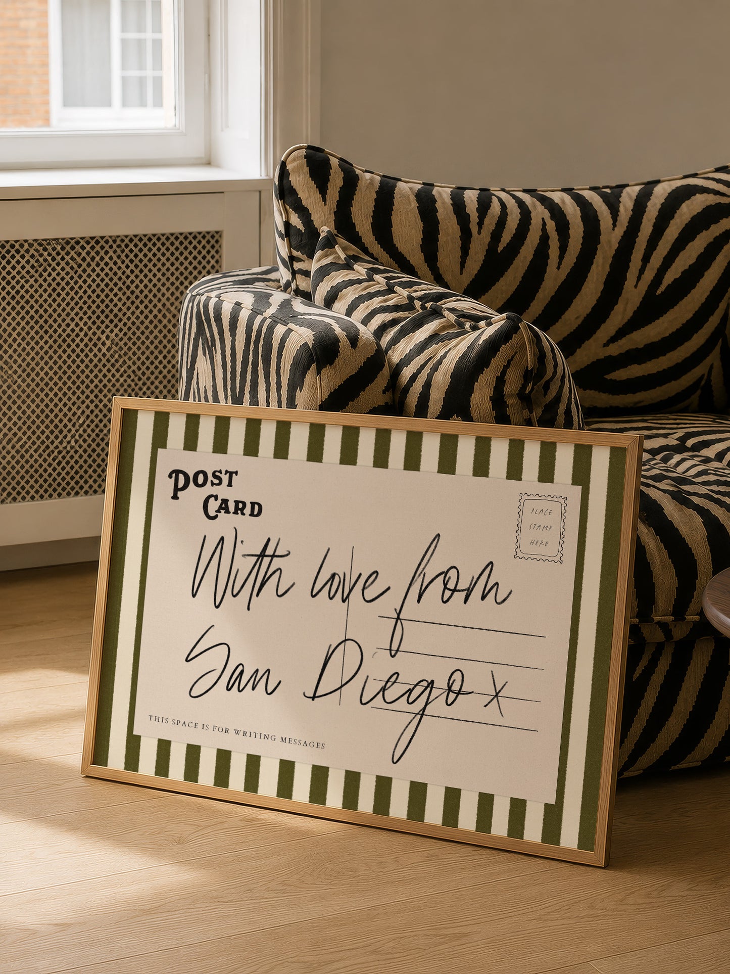 A framed postcard with a handwritten message is placed on a couch in a room with a window.