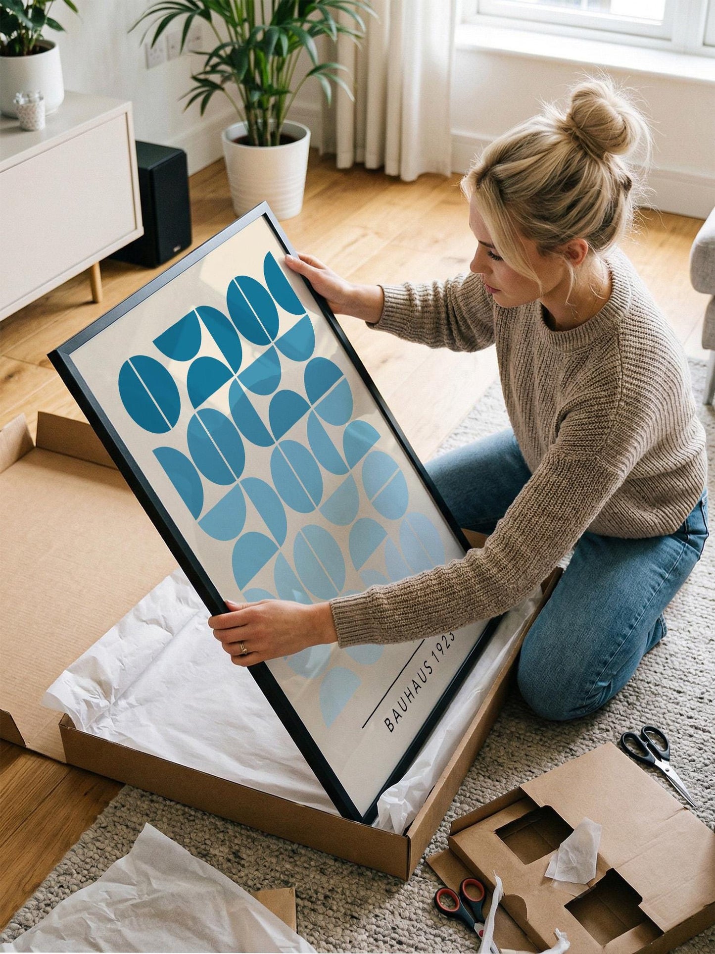A woman is kneeling on the floor and carefully opening a large picture frame, which is a Bauhaus-style artwork.