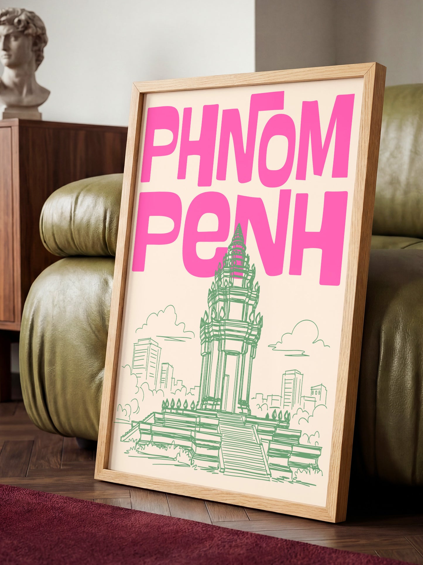 A framed poster with the text "PHNOM PENH" in pink and green, depicting a temple or monument, is displayed on a wooden floor in a room with a green couch.