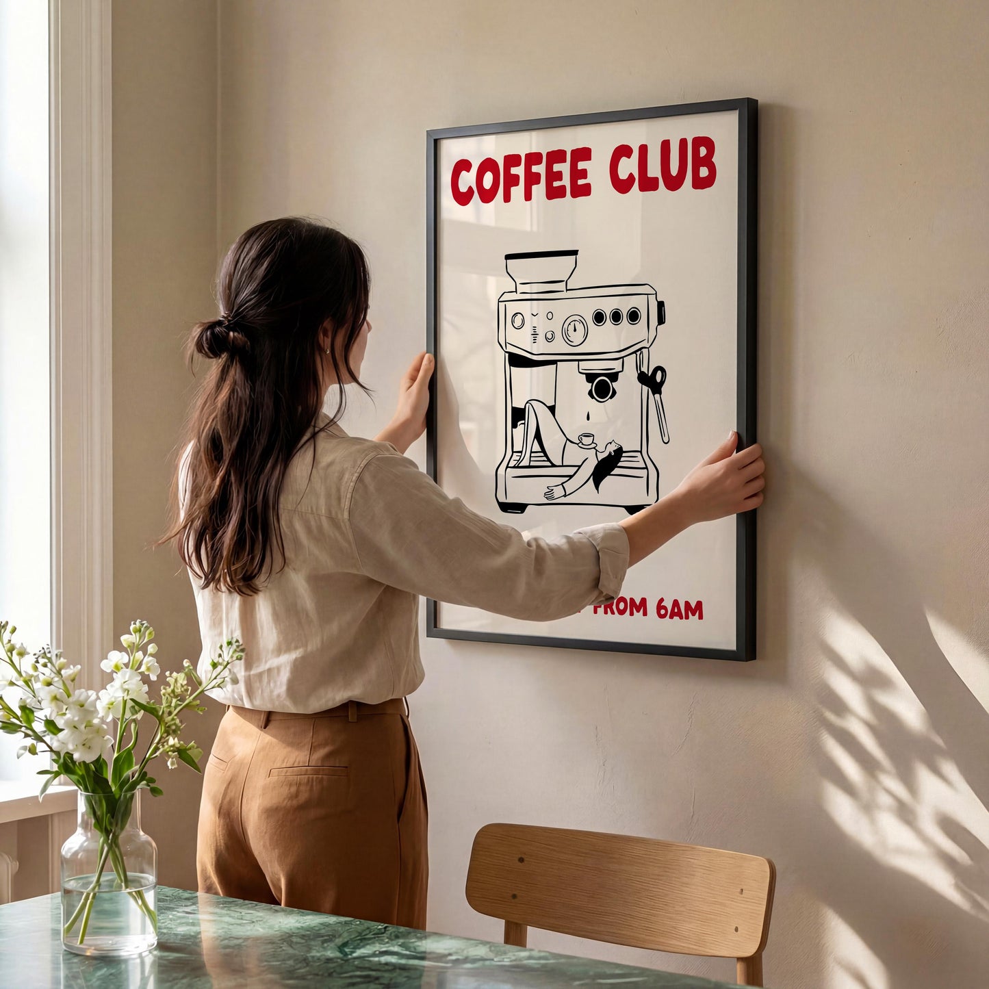 A woman hangs a framed poster on a wall, featuring a coffee machine and the text "COFFEE CLUB" in red letters.