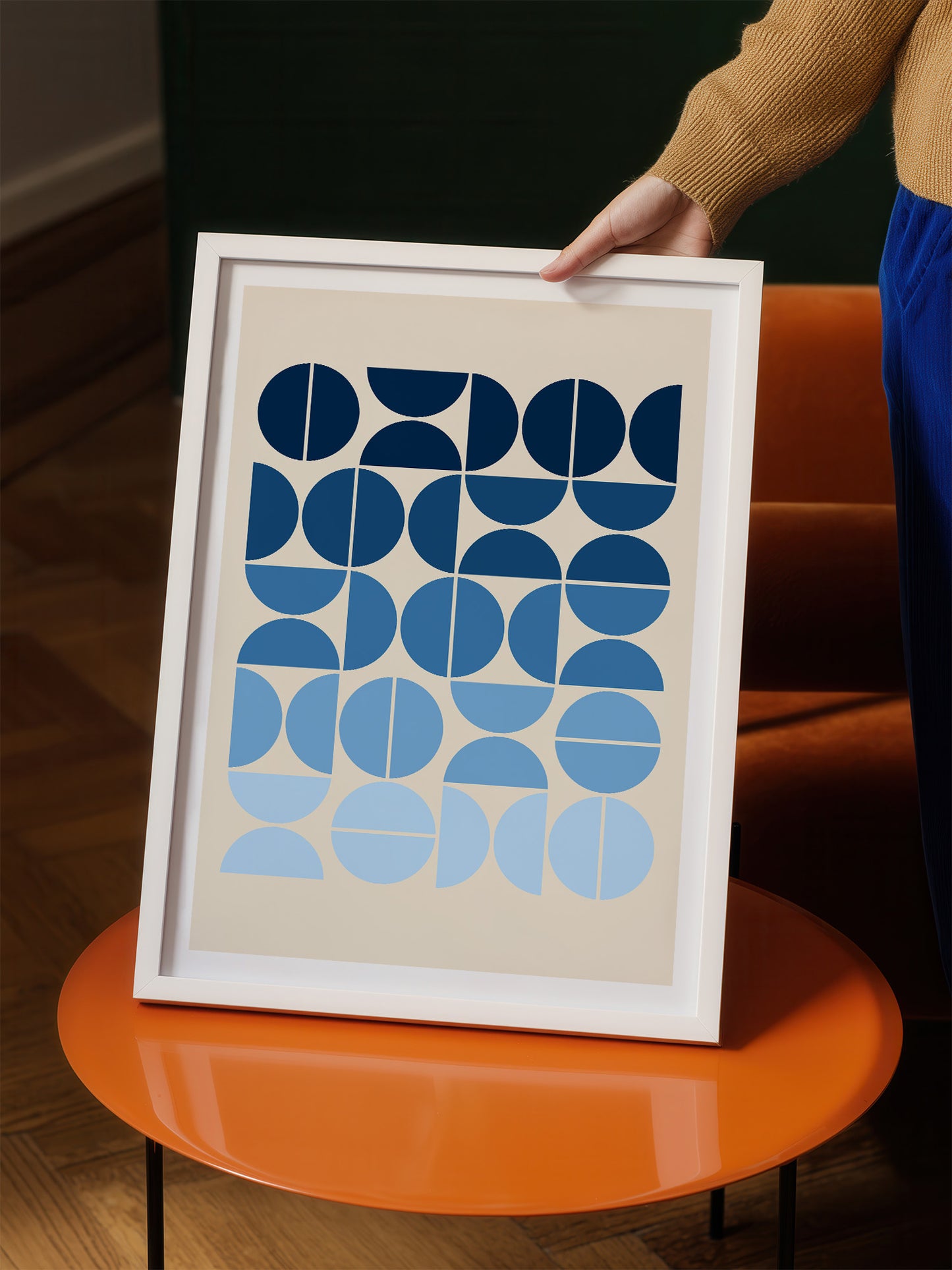 A person is holding up a framed print with a geometric design featuring blue circles and lines on a beige background.