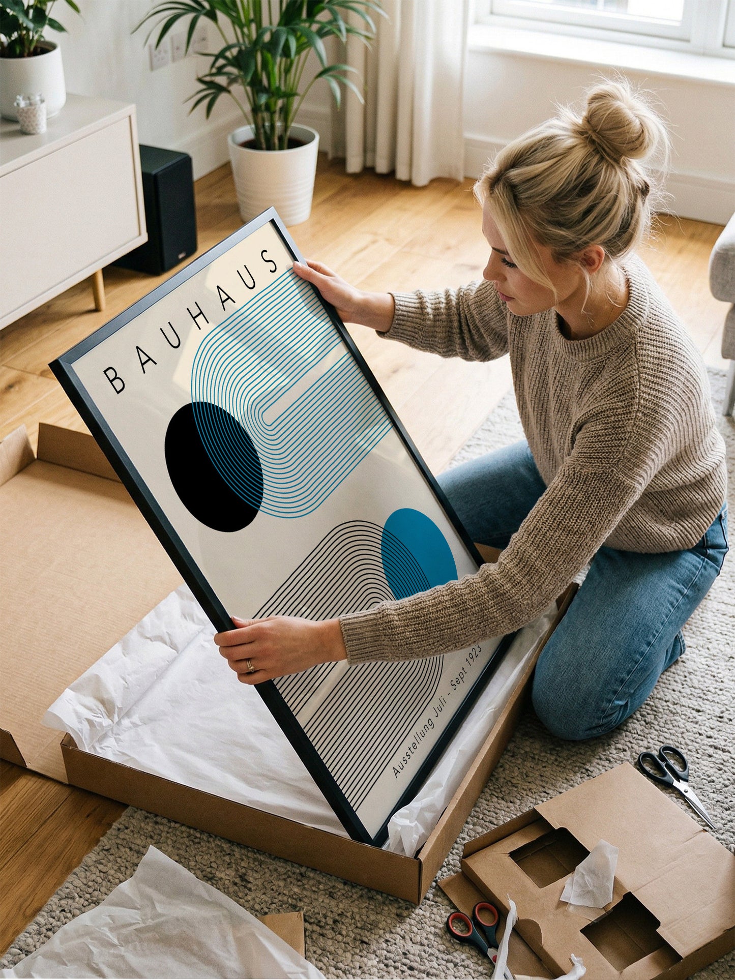 A woman is kneeling on the floor and holding a framed print of the Bauhaus art style.