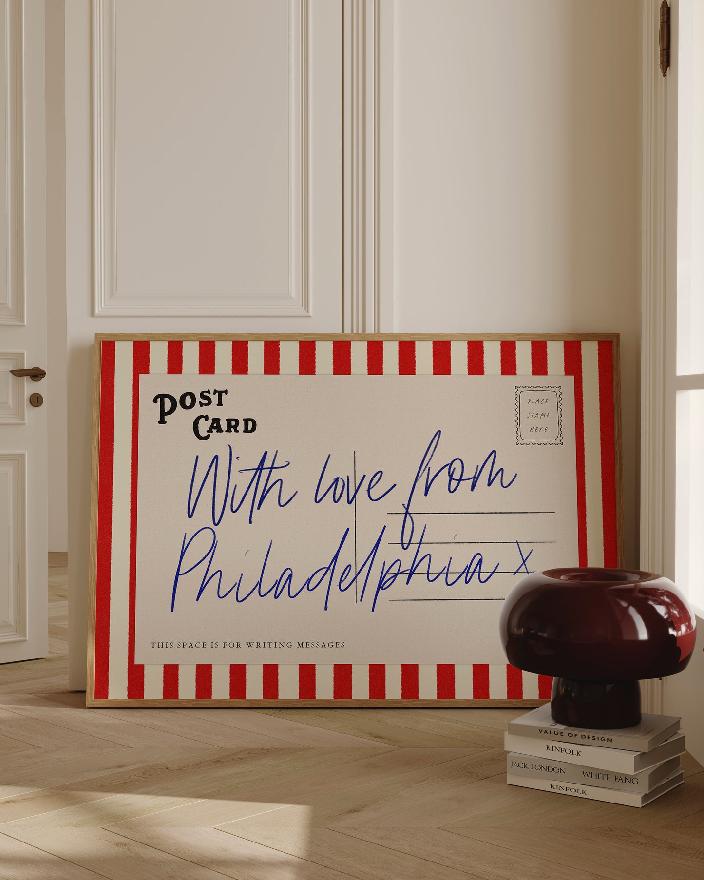 a large, red and white striped postcard with handwritten text on it, placed on a wooden floor next to a stack of books.