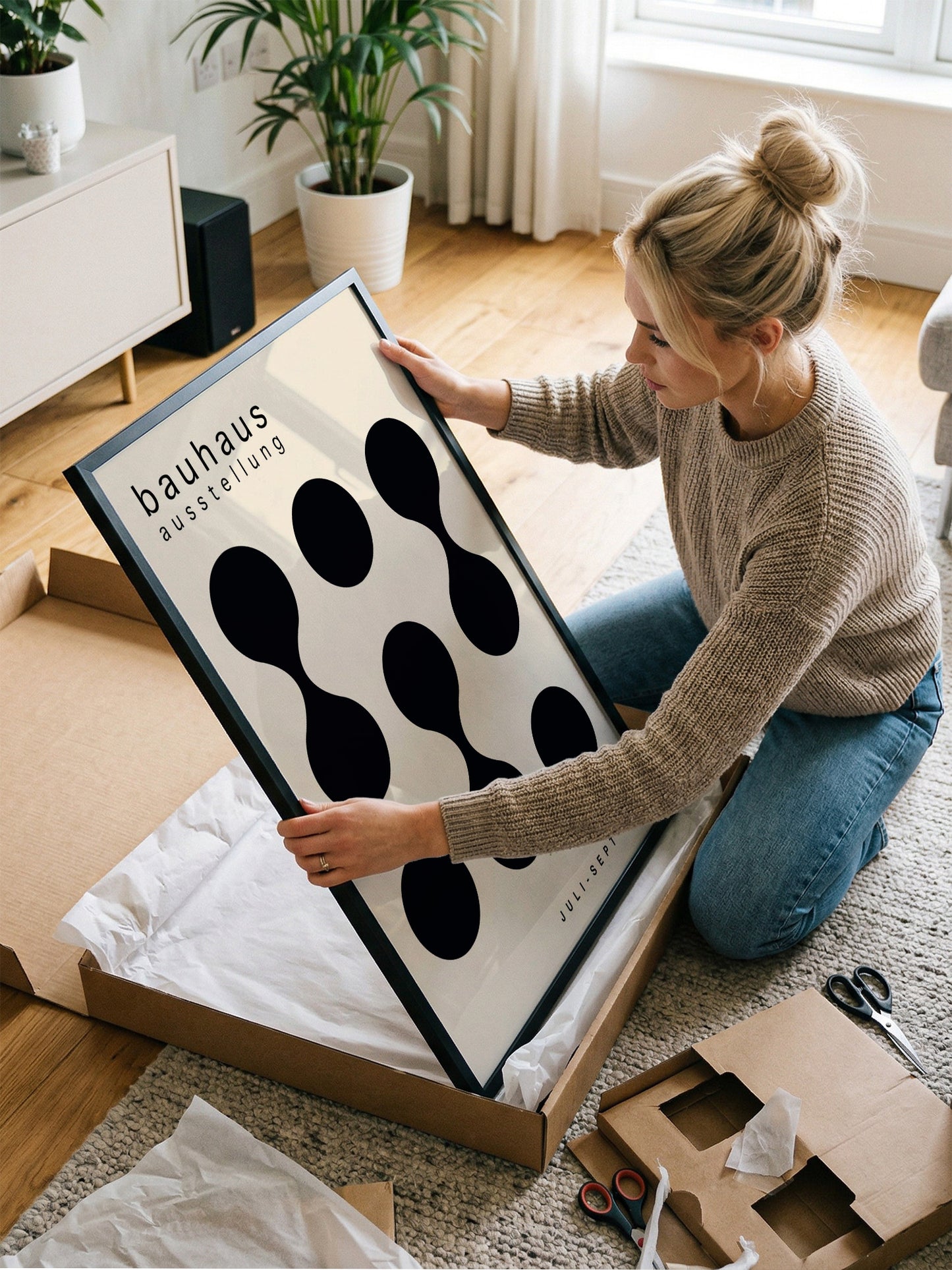 A woman is kneeling on the floor and holding a large framed print with a black and white design.