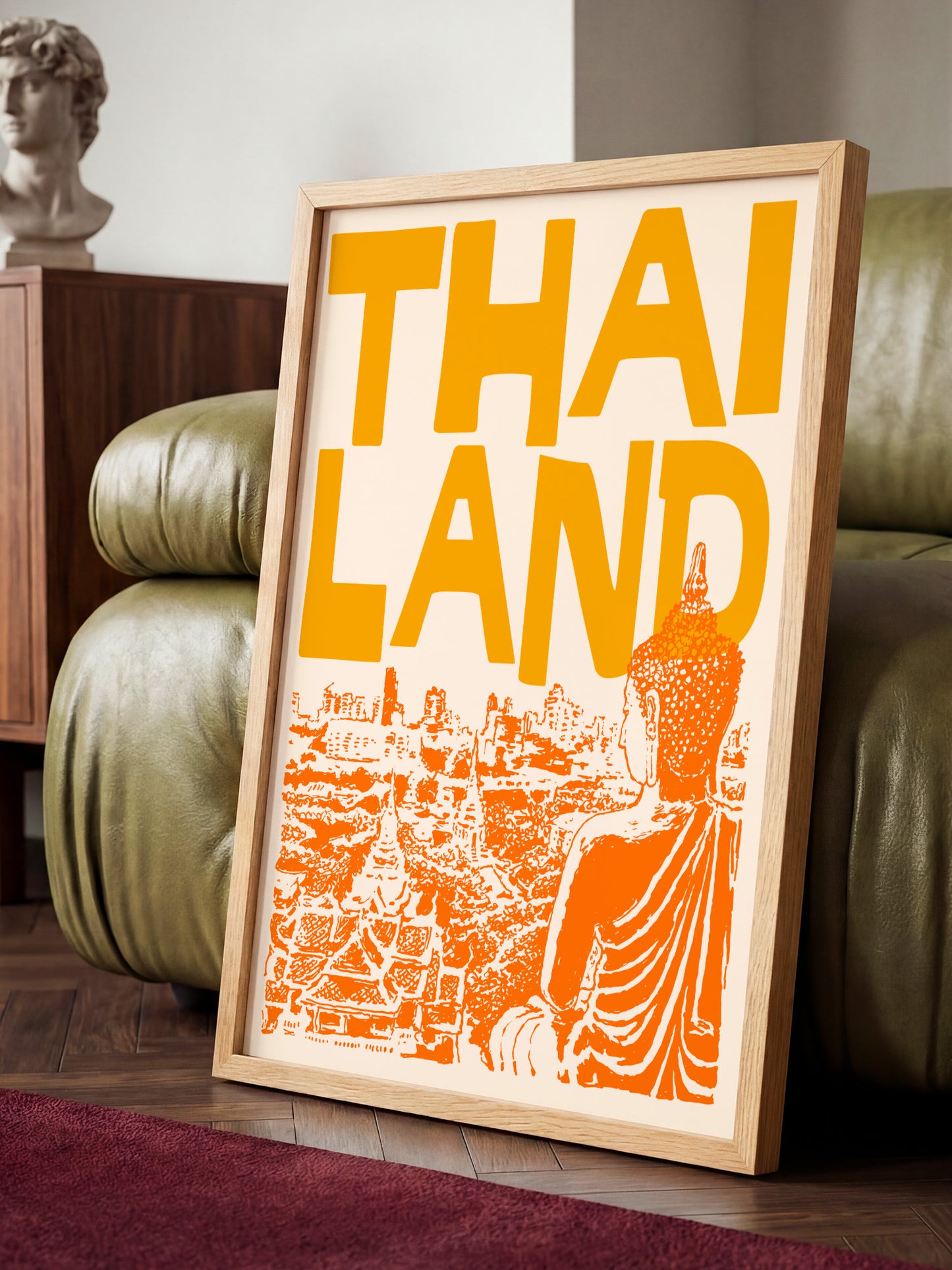 A framed poster with the text "THAI LANDS" and an illustration of Buddha in an orange robe, placed on a wooden floor in a room with a green couch.