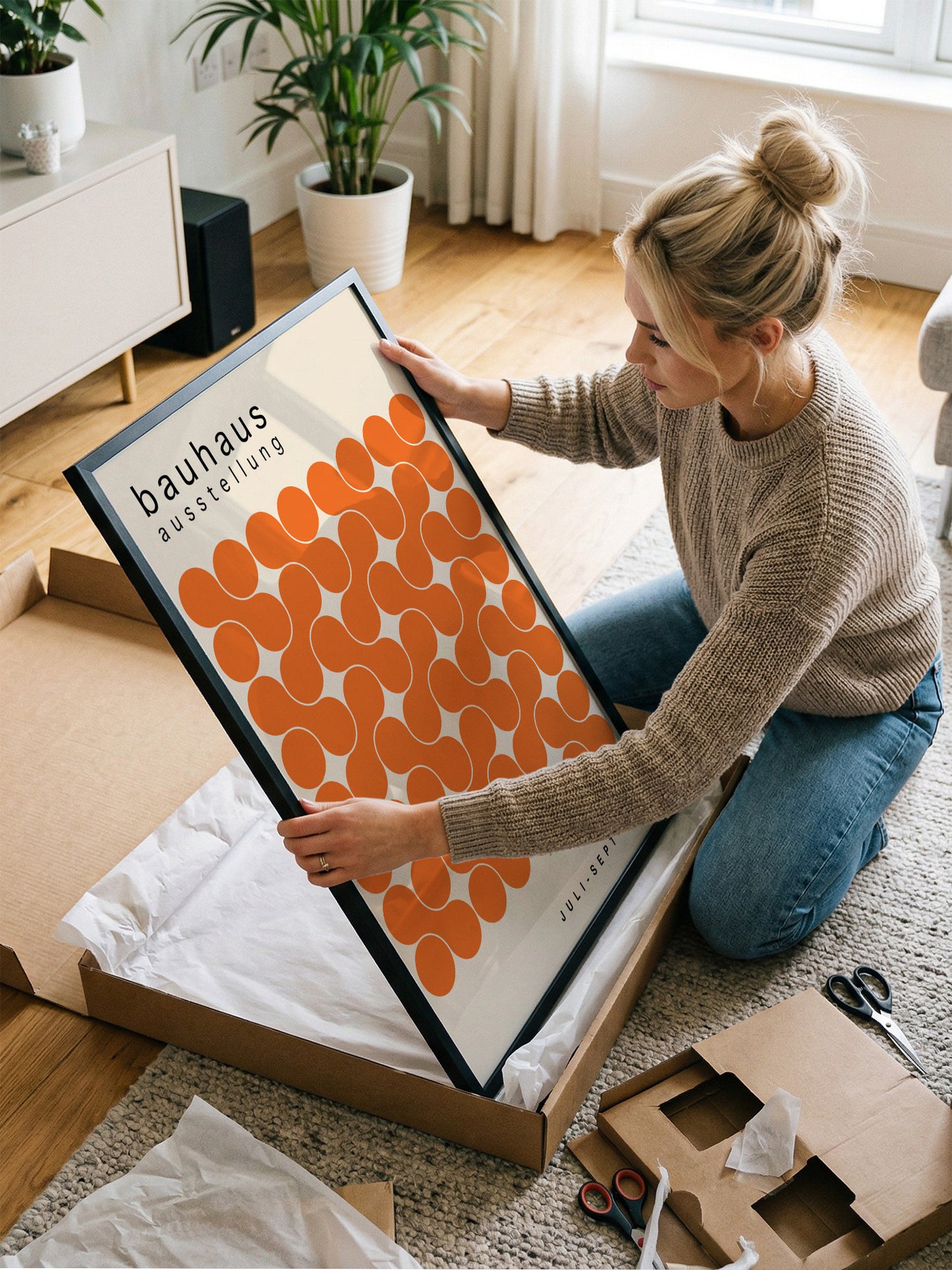 A woman is kneeling on the floor and holding a framed print with an orange and white design.